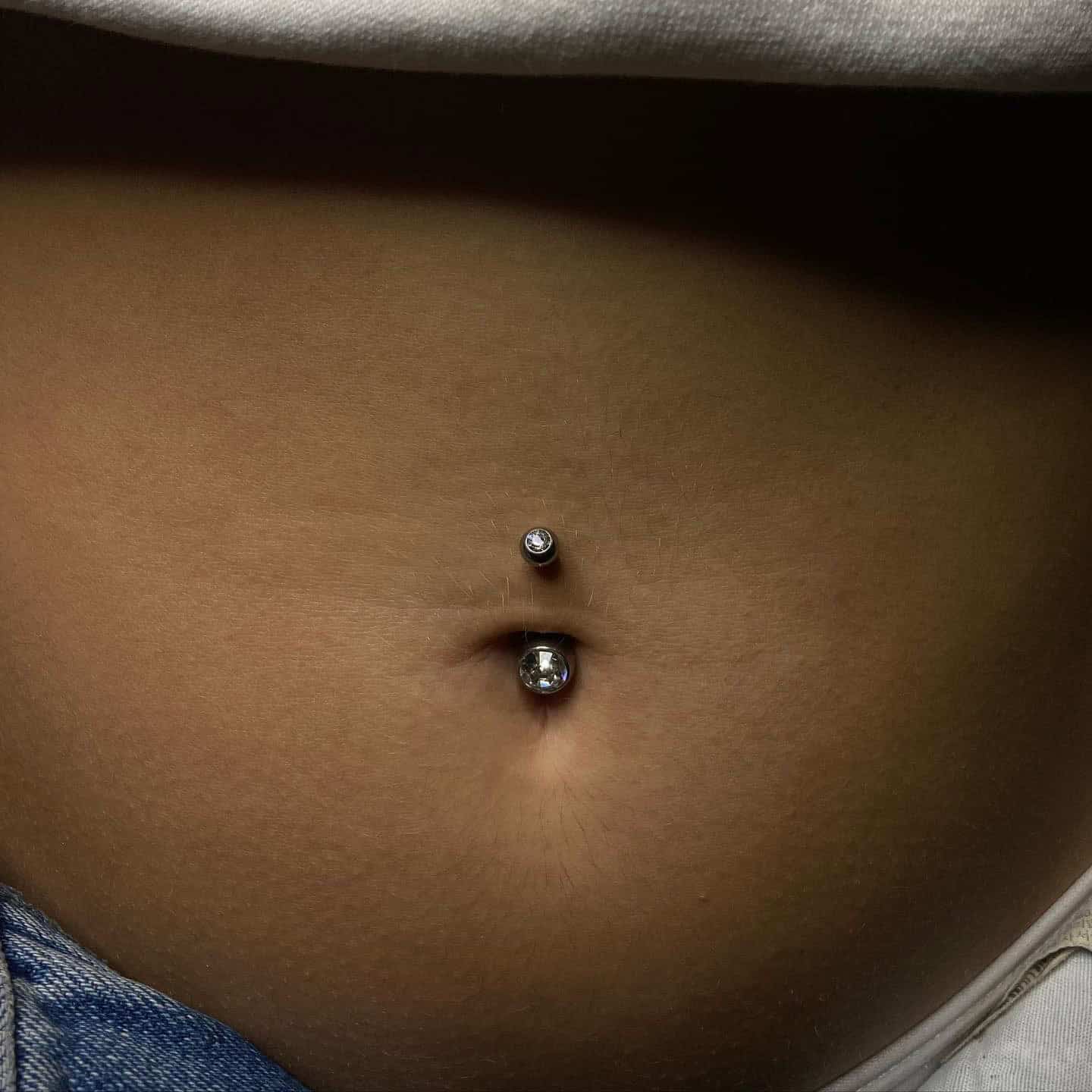 7sinstattoo.live Zbliżenie pępka osoby ozdobionego piercingiem 01E732A3-9C82-4252-8736-0EEBB9E24FD4. Piercing ma srebrny ćwiek z przezroczystym kamieniem szlachetnym w górnej części i większą srebrną kulkę z przezroczystym kamieniem szlachetnym w dolnej części. Osoba ma na sobie jasny top i niebieskie dżinsy, idealne na wizytę w studio tatuażu Poznań. Najlepsze studio tatuażu Seven Sins Tattoo