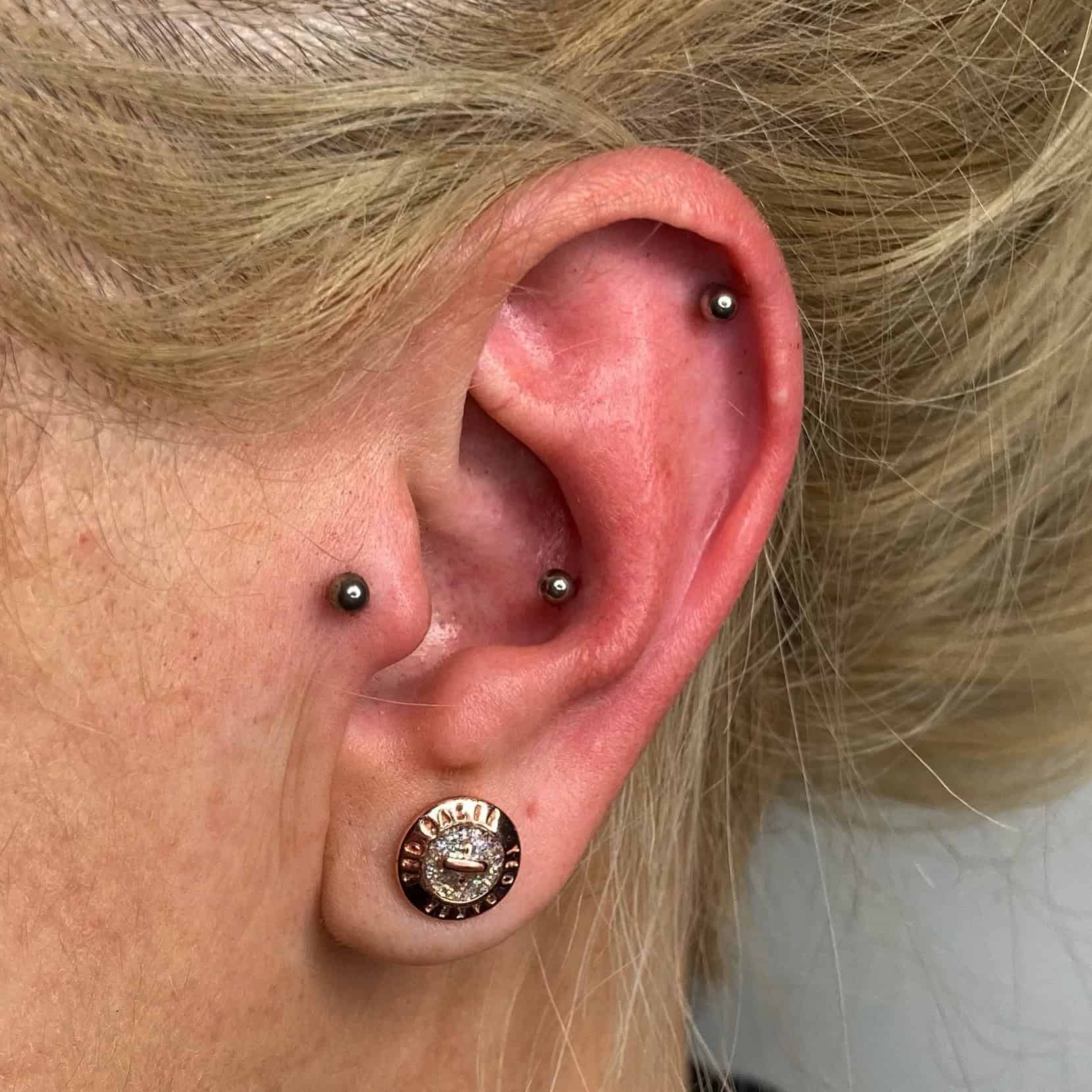 7sinstattoo.live Zbliżenie ucha osoby z trzema piercingami, zrobione w Studio Tatuażu Poznań. Płatek ucha ozdobiony jest kolczykami 04790D2D-A355-4DFA-94C3-8EFFB7822948 z okrągłym wzorem z tekstem i centralnym emblematem. Obszar chrząstki ma dwa mniejsze okrągłe ćwieki, jeden na helisie, a drugi w wewnętrznej części muszli. Osoba ma jasnoblond włosy. Najlepsze studio tatuażu Seven Sins Tattoo