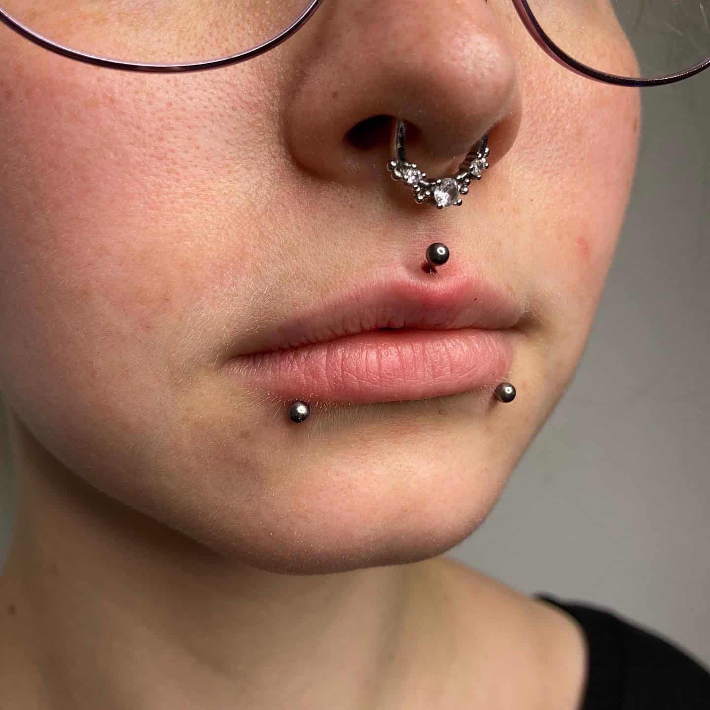 7sinstattoo.live Zbliżenie dolnej części twarzy osoby, prezentujące jej misterne piercingi twarzy, zostało pięknie uchwycone w studio tatuażu Poznań. Osoba jest ozdobiona piercingiem przegrody nosowej o misternym wzorze i trzema piercingami warg: jednym umieszczonym centralnie pod dolną wargą i dwoma po obu stronach nad brodą. Nosi również okulary. Ten obraz jest zidentyfikowany jako Nazwa produktu 06219259-71A6-4A73-B889-7A84A896FAC3. Najlepsze studio tatuażu Seven Sins Tattoo