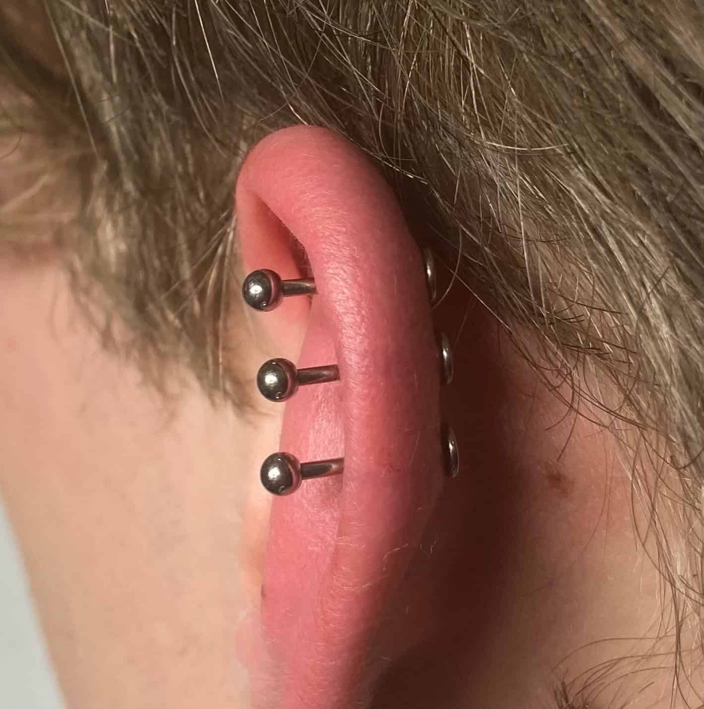 7sinstattoo.live Zbliżenie ucha przekłutego trzema przemysłowymi sztangami 0B2BB22E-0D99-4030-A1D5-28F5857EDCF6, z których każda ma małe srebrne kulki na obu końcach. Ucho ma czerwonawe odcienie wokół przekłuć, na tle jasnobrązowych włosów. Zrobione w Studio Tatuaże Poznań, znanym z wyjątkowej dbałości o szczegóły. Najlepsze studio tatuażu Seven Sins Tattoo