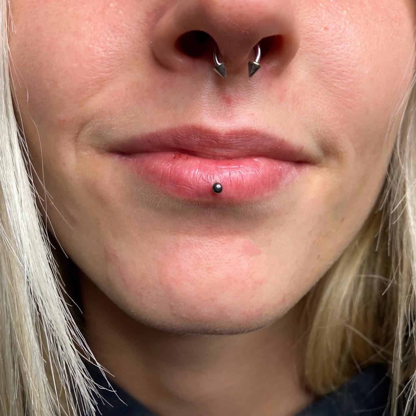 7sinstattoo.live Zbliżenie ust i nosa osoby ukazuje piercing przegrody nosowej z podkową ozdobioną dwoma kolcami, obok piercingu wargi dolnej z małą kulką. Osoba ma jasną cerę i blond włosy, charakterystyczne dla klientów studia tatuażu Poznań, prezentującego produkt 16924757-9863-467A-A3B5-E3B45A85F898. Najlepsze studio tatuażu Seven Sins Tattoo