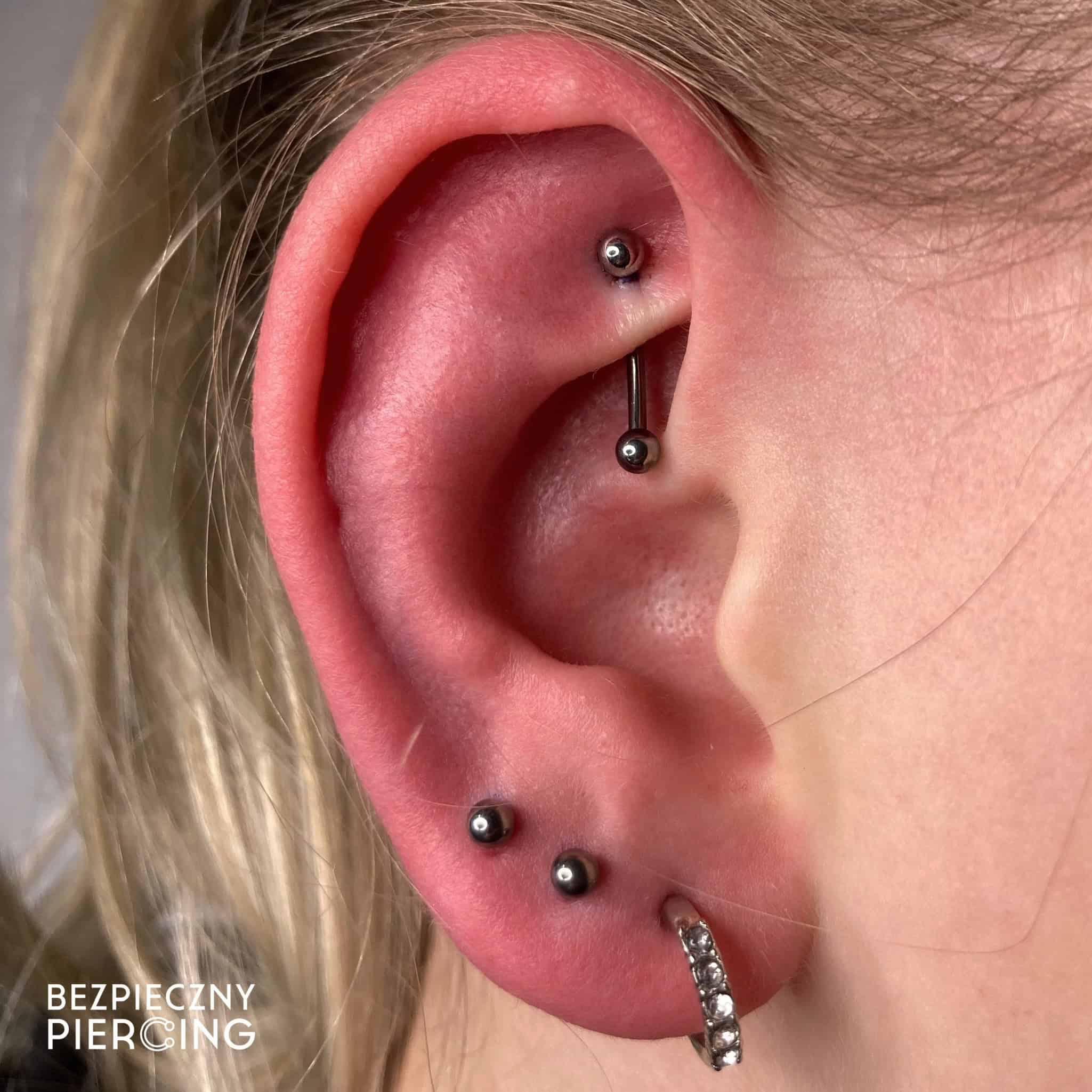 7sinstattoo.live Zbliżenie ucha osoby ozdobionego trzema różnymi piercingami: sztangą w chrząstce, dwoma kolczykami w płatku i kółkiem wysadzanym kamieniami szlachetnymi w dolnym płatku. Obraz zawiera tekst w lewym dolnym rogu, który brzmi „BEZPIECZNY PIERCING” ze studia tatuaży Poznań. Polecany produkt: 17A15EDF-8872-4FA1-9B55-F0A3AE14C62B. Najlepsze studio tatuażu Seven Sins Tattoo