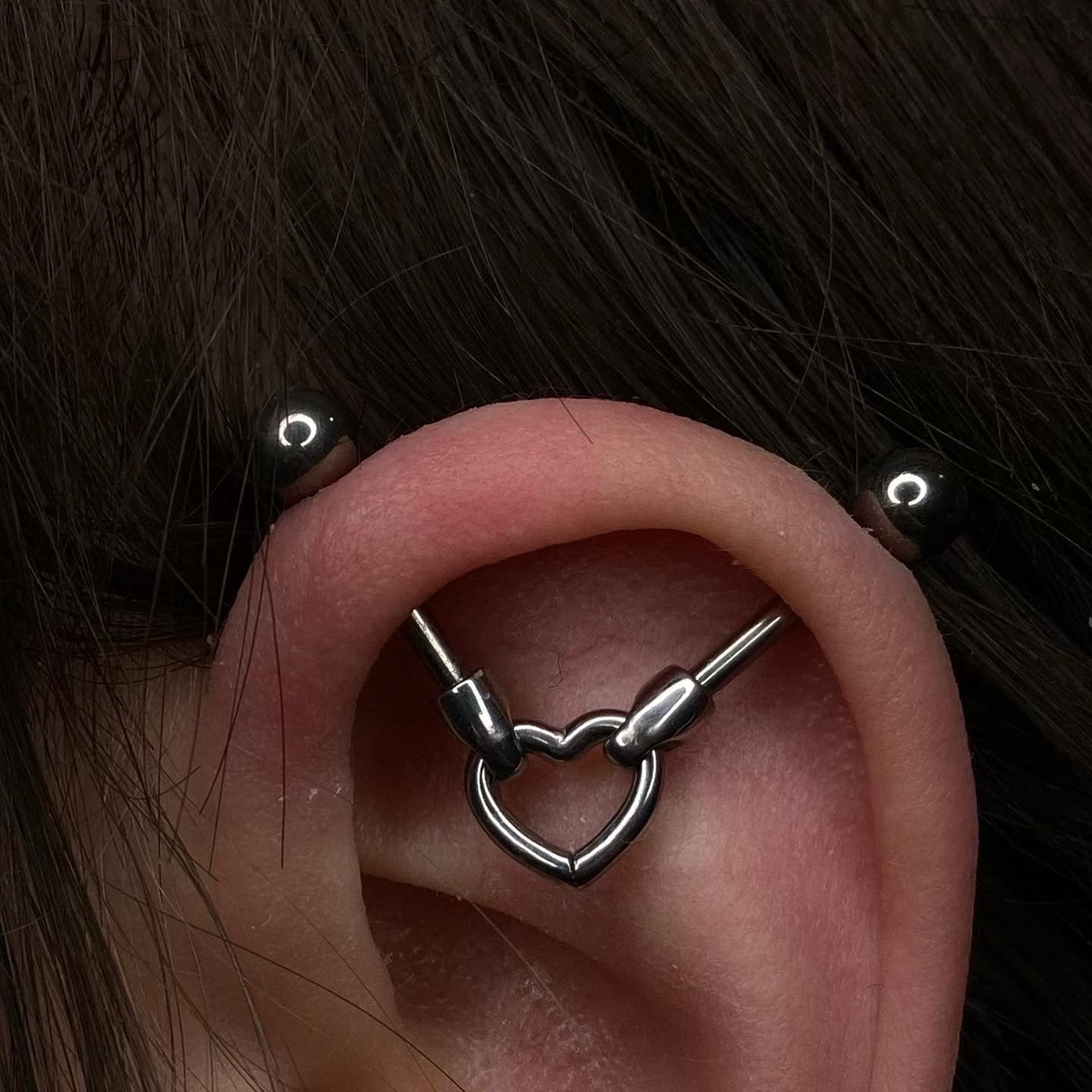 7sinstattoo.live Zbliżenie ucha z industrialnym piercingiem. Kolczyk ma dwa końce, które łączą się z metalowym środkiem w kształcie serca na środku ucha. Biżuteria z produktu 1DCFB785-E9DE-49EC-84CC-3B1133C71388 jest osadzona na tle ciemnych włosów, eksponując misterne dzieło, które można znaleźć w studio tatuażu Poznań. Najlepsze studio tatuażu Seven Sins Tattoo