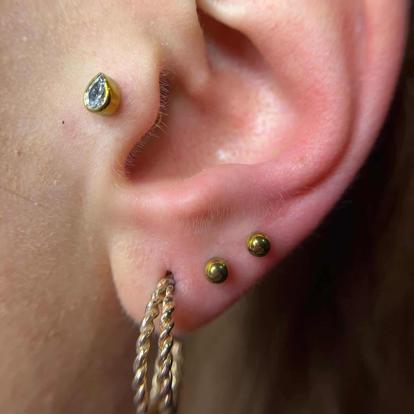 7sinstattoo.live Zbliżenie ucha z trzema piercingami, zrobione w studio tatuażu Poznań. Na górnej części ucha znajduje się mały kolczyk z kamieniem szlachetnym, na dolnej dwa małe złote kolczyki w kształcie kulek, a na płatku ucha gruby, skręcony złoty kolczyk-kółko. Wszystkie prezentowane kolczyki pochodzą z produktu 2057CC28-B589-4B9C-9277-4E72C18439B6. Najlepsze studio tatuażu Seven Sins Tattoo