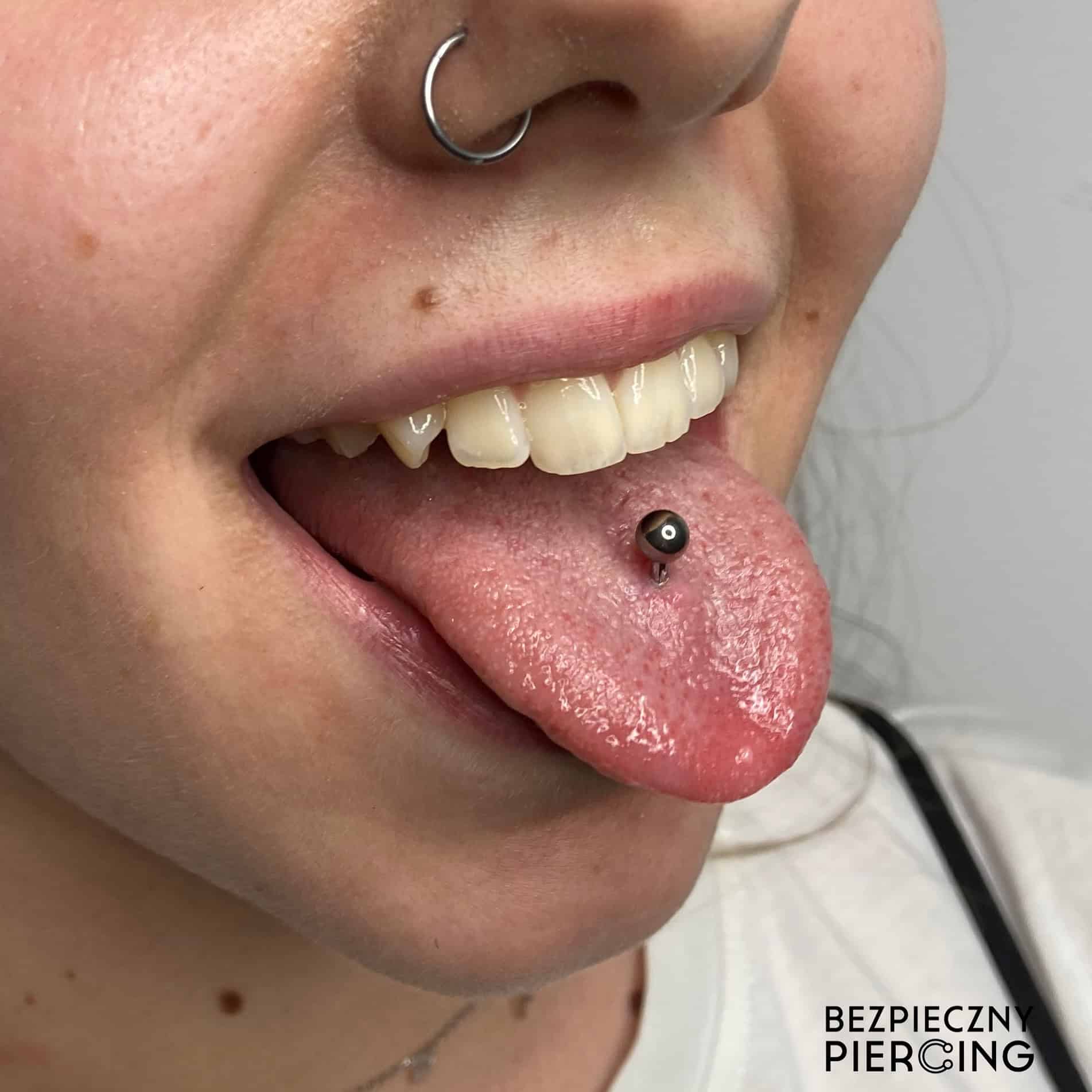 7sinstattoo.live Zbliżenie twarzy osoby ukazuje kolczyk w lewym nozdrzu i piercing języka ozdobiony metalowym sztangą. Osoba uśmiecha się z otwartymi ustami, eksponując białe zęby. W prawym dolnym rogu tekst „BEZPIECZNY PIERCING” podkreśla kompetencje naszego poznańskiego studia tatuażu, prezentując produkt 23B5387E-E272-4E64-AB43-AE5070EC0043. Najlepsze studio tatuażu Seven Sins Tattoo