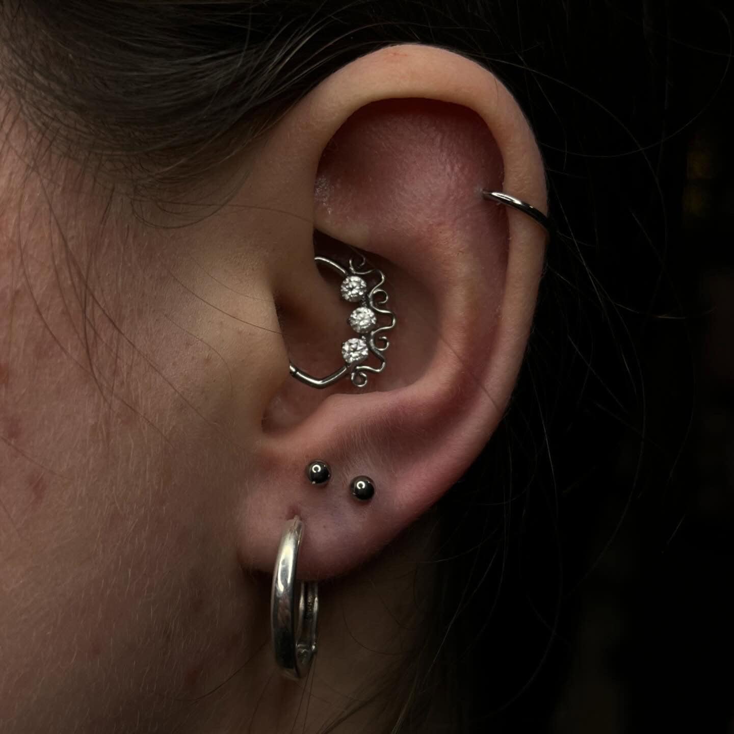 7sinstattoo.live Zbliżenie na ucho z wieloma piercingami w studio tatuażu Poznań. Zewnętrzna chrząstka ma kształt kolczyka-kółka 266BE792-3702-487A-858F-889F8893605F, wewnętrzna chrząstka ma zakrzywiony sztangę ozdobioną trzema błyszczącymi klejnotami, a dolne płatki są ozdobione dwoma małymi ćwiekami i kolejnym dużym kolczykiem-kółkiem. Najlepsze studio tatuażu Seven Sins Tattoo