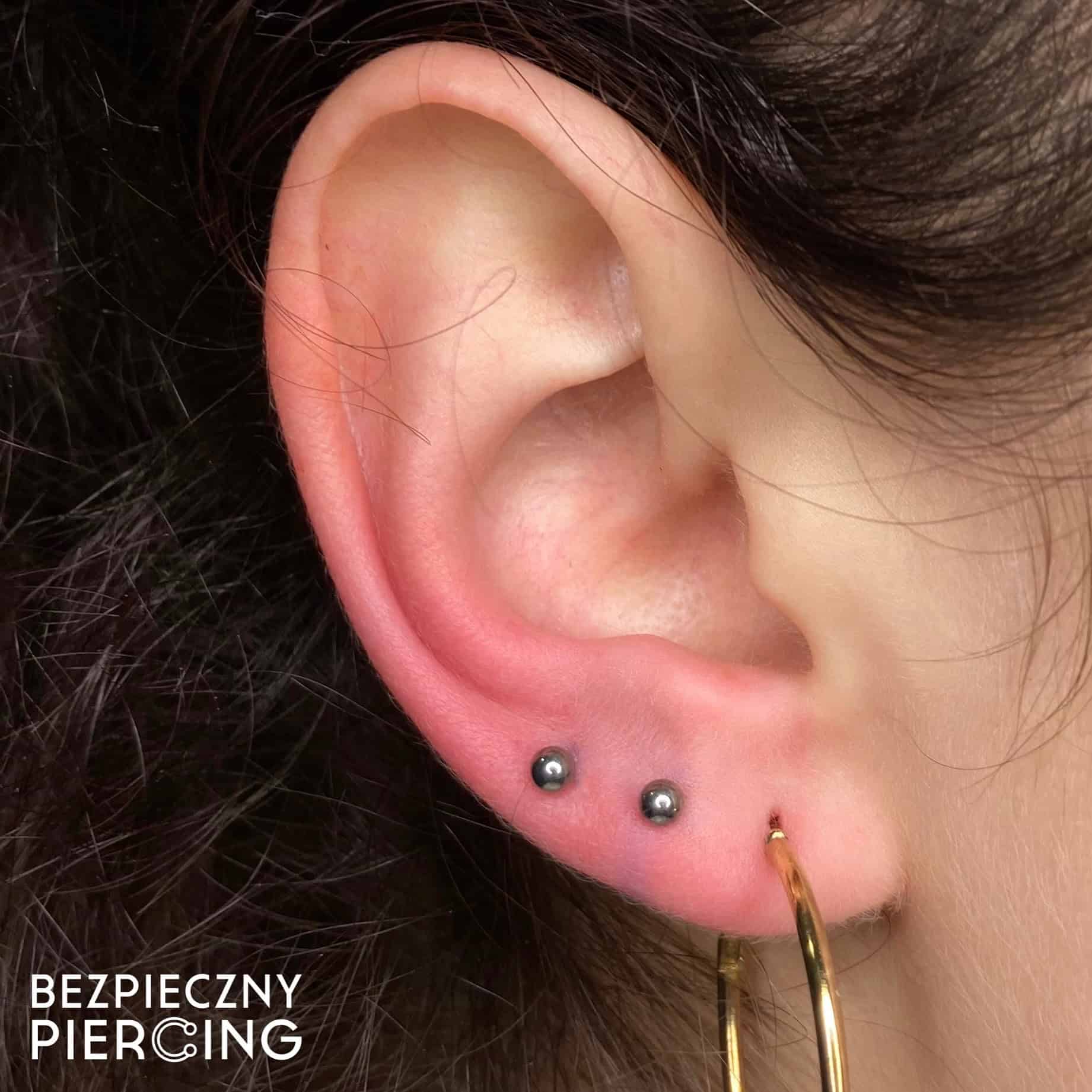 7sinstattoo.live Zbliżenie na ucho osoby z dwoma małymi czarnymi kolczykami na płatku i dużym złotym kolczykiem w kształcie koła w dolnym płatku. W tle widać ciemne włosy, a w lewym dolnym rogu wyświetlany jest tekst „BEZPIECZNY PIERCING”, podświetlający produkt 27664927-1D55-4BE8-BE0E-260FE4AFE6BE z naszego studia tatuaży Poznań. Najlepsze studio tatuażu Seven Sins Tattoo