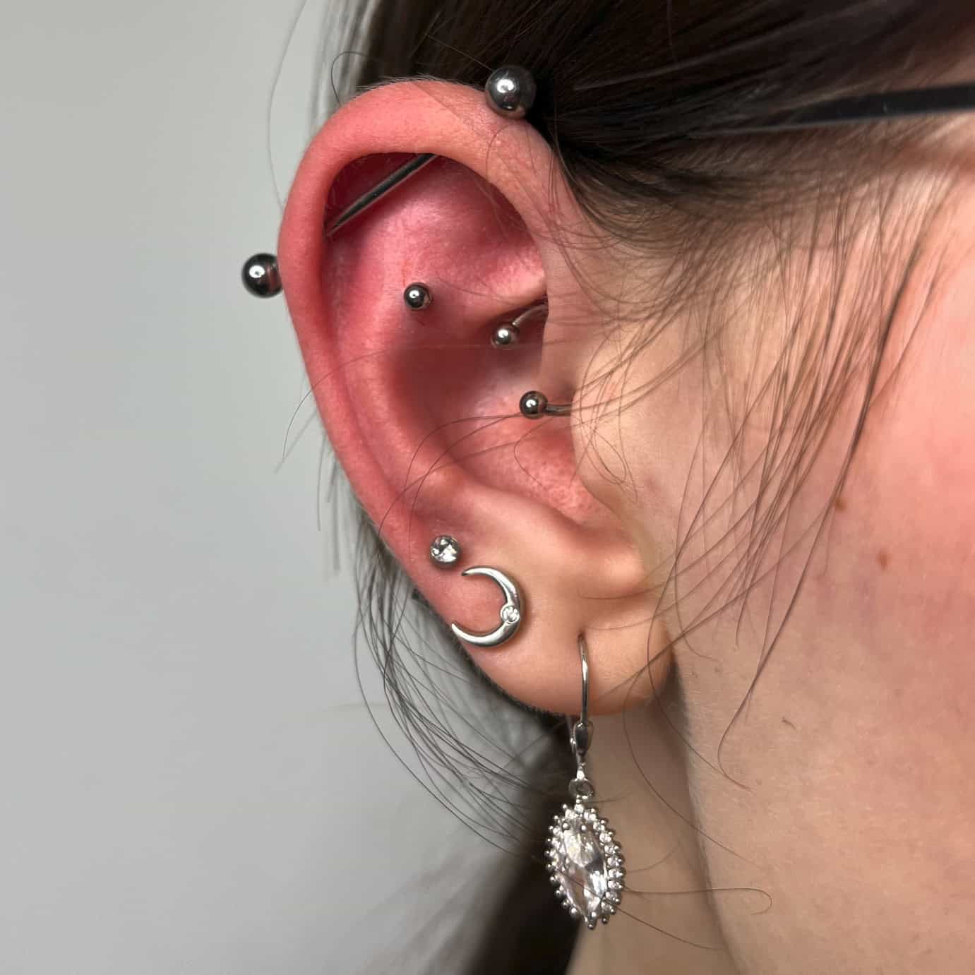 7sinstattoo.live Zbliżenie ucha osoby ozdobionego wieloma piercingami w najlepszym studiu tatuażu w Poznaniu. Ucho ma mieszankę biżuterii, w tym kolczyki, małe kółko z zawieszką w kształcie półksiężyca i zwisający kolczyk z ozdobnym wzorem i dużym, przezroczystym kamieniem szlachetnym z kolekcji **32729F66-76D4-4F28-8B98-382AE417F714**. Włosy osoby są schowane za uchem. Najlepsze studio tatuażu Seven Sins Tattoo