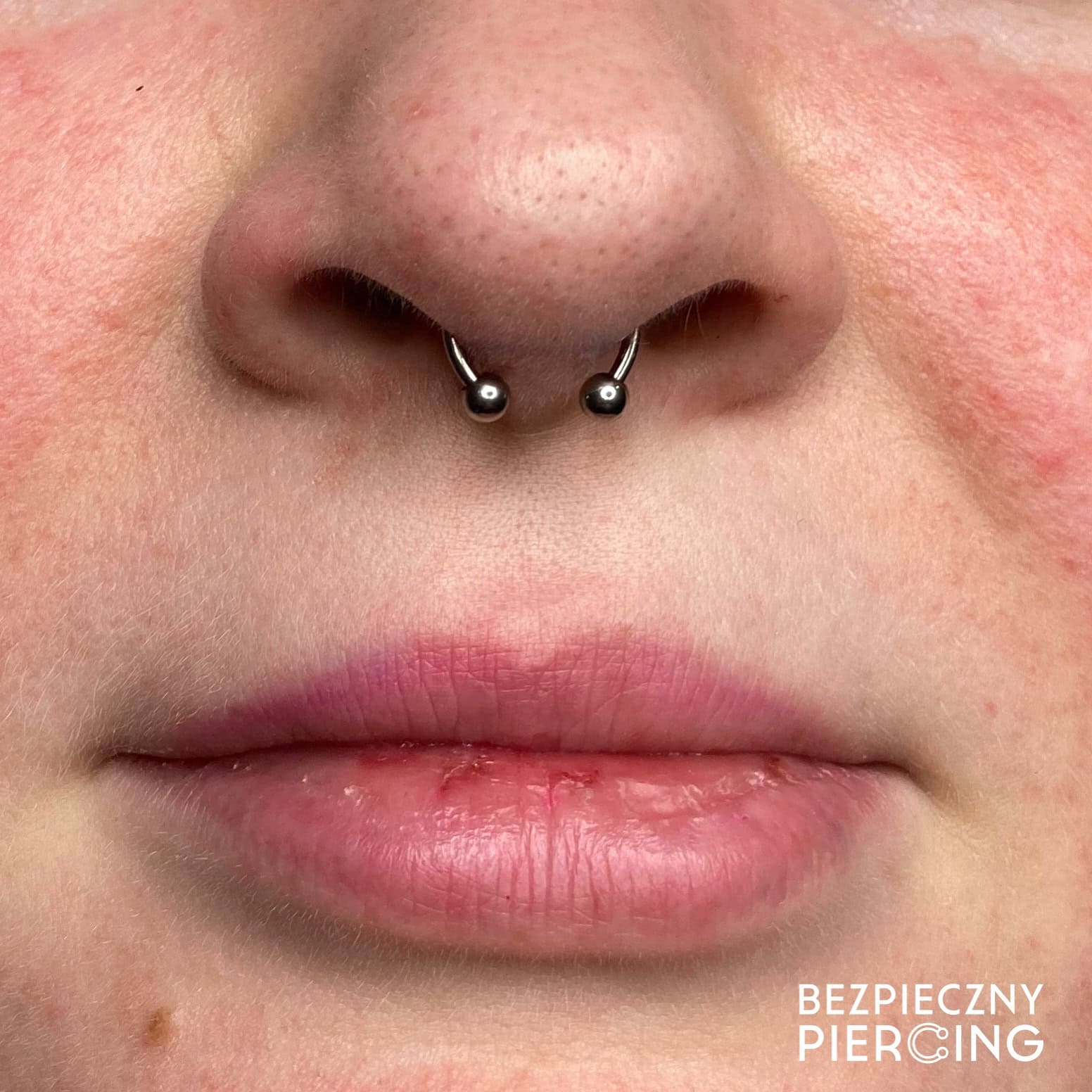 7sinstattoo.live Zbliżenie dolnej części twarzy osoby, skupiające się na jej ustach i nosie, prezentujące piercing przegrody nosowej ozdobiony produktem 3C85E30B-74B7-446B-9610-4EEAC719D190 - kółkiem w kształcie podkowy z dwoma małymi kulkami na końcach. Skóra ma naturalną fakturę i kilka piegów. W prawym dolnym rogu pojawia się napis „BEZPIECZNY PIERCING”, wykonany przez najlepsze studio tatuażu Poznań. Najlepsze studio tatuażu Seven Sins Tattoo