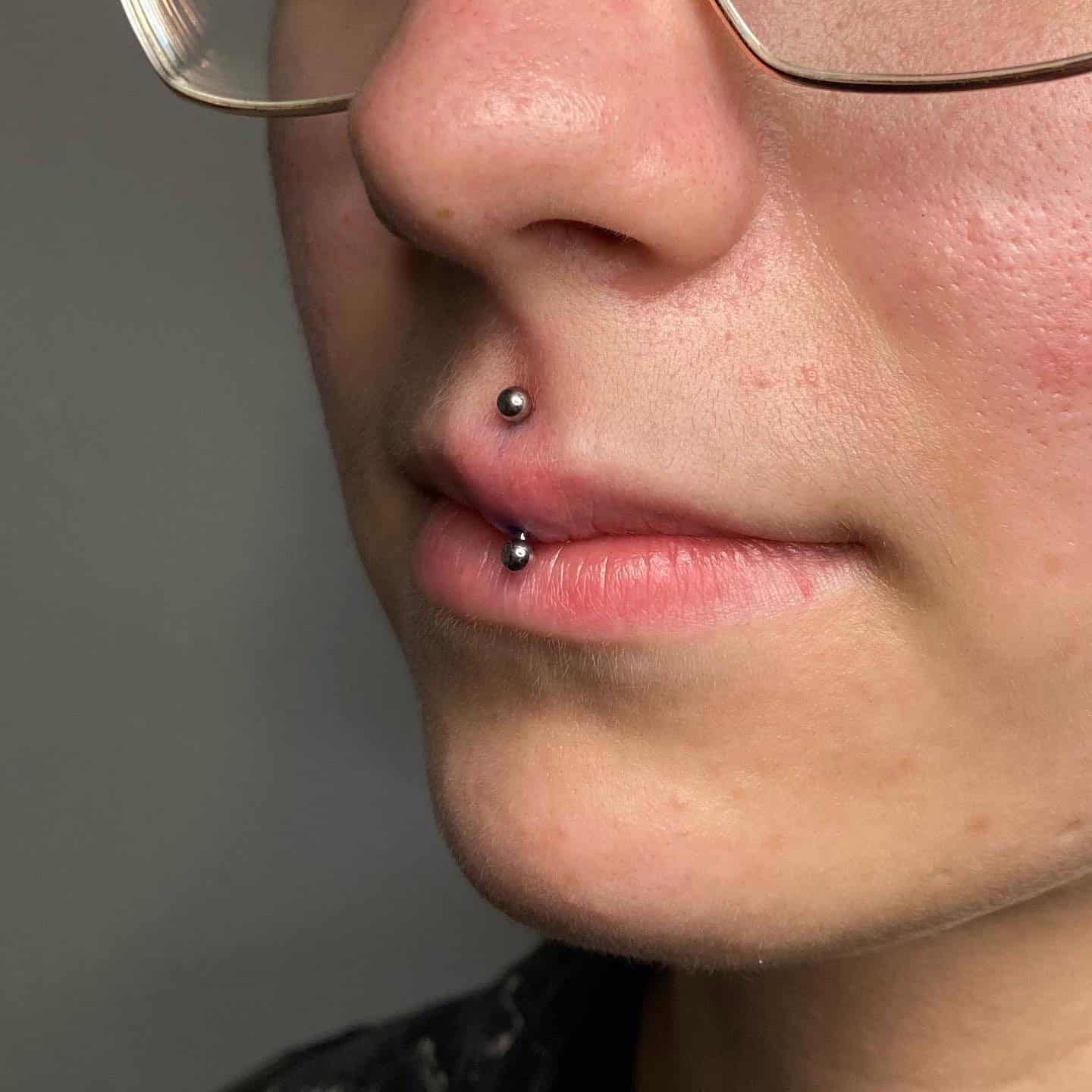 7sinstattoo.live Zbliżenie twarzy osoby noszącej okulary, z pionowym piercingiem wargi górnej ozdobionym jednym koralikiem nad górną wargą i drugim pod dolną wargą ze studia tatuażu Poznań. Tło jest gładkie i nieostre. Produkt użyty do piercingu jest zidentyfikowany jako 3CCABBAA-8141-4D1B-947C-B3DD1DF3D99F. Najlepsze studio tatuażu Seven Sins Tattoo