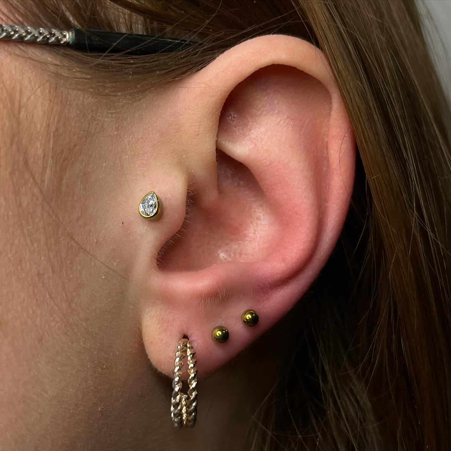 7sinstattoo.live Zbliżenie ucha osoby pokazujące różne kolczyki, w tym produkt 409414AB-0BA3-4CBA-9D6A-3D1B33ECAE16. W górnej chrząstce znajduje się gruszkowaty diamentowy kolczyk, dwa małe złote kolczyki wzdłuż zewnętrznej części ucha i teksturowana złota obręcz w płatku ucha. Osoba nosi również okulary z czarno-złotymi oprawkami, stylowo uchwycone w study tatuażu Poznań. Najlepsze studio tatuażu Seven Sins Tattoo