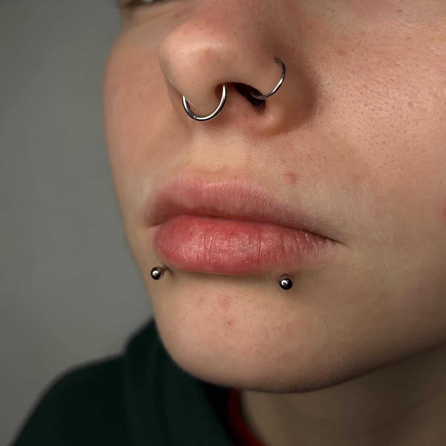 7sinstattoo.live Zbliżenie dolnej części twarzy osoby pokazujące dwa piercingi wargi z małymi, czarnymi koralikami po obu stronach dolnej wargi i piercing przegrody nosowej z kółkiem w kształcie podkowy. Osoba ma neutralny wyraz twarzy, a część ciemnej koszuli jest widoczna. Zrobione w jednym z najlepszych studiów tatuaży w Poznaniu. Prezentuje produkt 410E2558-F30F-417D-B386-375B79E61F1C. Najlepsze studio tatuażu Seven Sins Tattoo