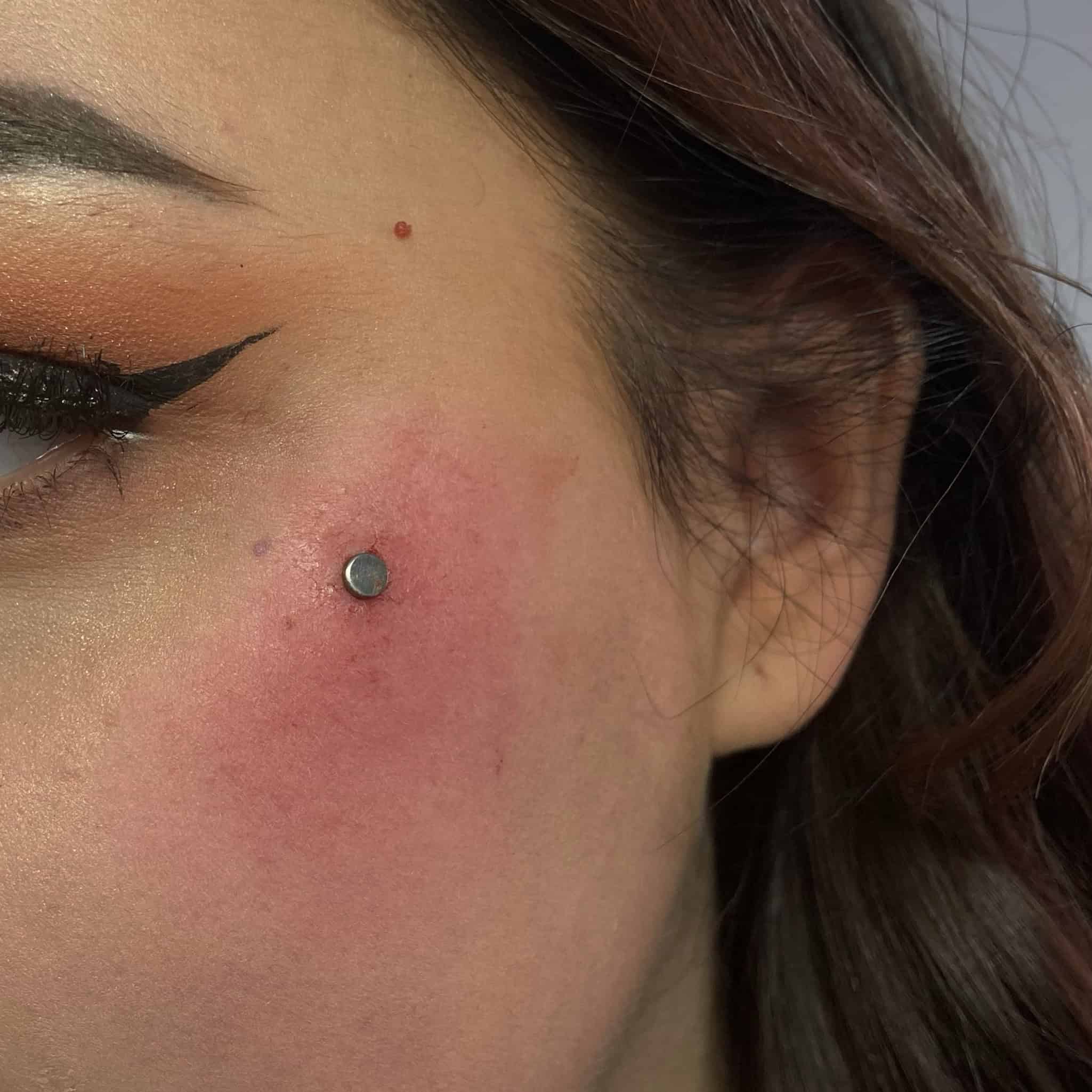 7sinstattoo.live Zbliżenie lewej strony twarzy kobiety, prezentujące świeży piercing policzka z okrągłym, srebrnym ćwiekiem ozdobionym produktem 4468FFAC-CA96-4AEA-966D-A3FCD9115568. Przekłuty obszar wydaje się lekko zaczerwieniony i opuchnięty. Ma skrzydlatą kreskę na powiece i lekko nałożony makijaż, a jej ciemnobrązowe włosy są częściowo widoczne — doskonały przykład z najlepszego studia tatuażu Poznań. Najlepsze studio tatuażu Seven Sins Tattoo