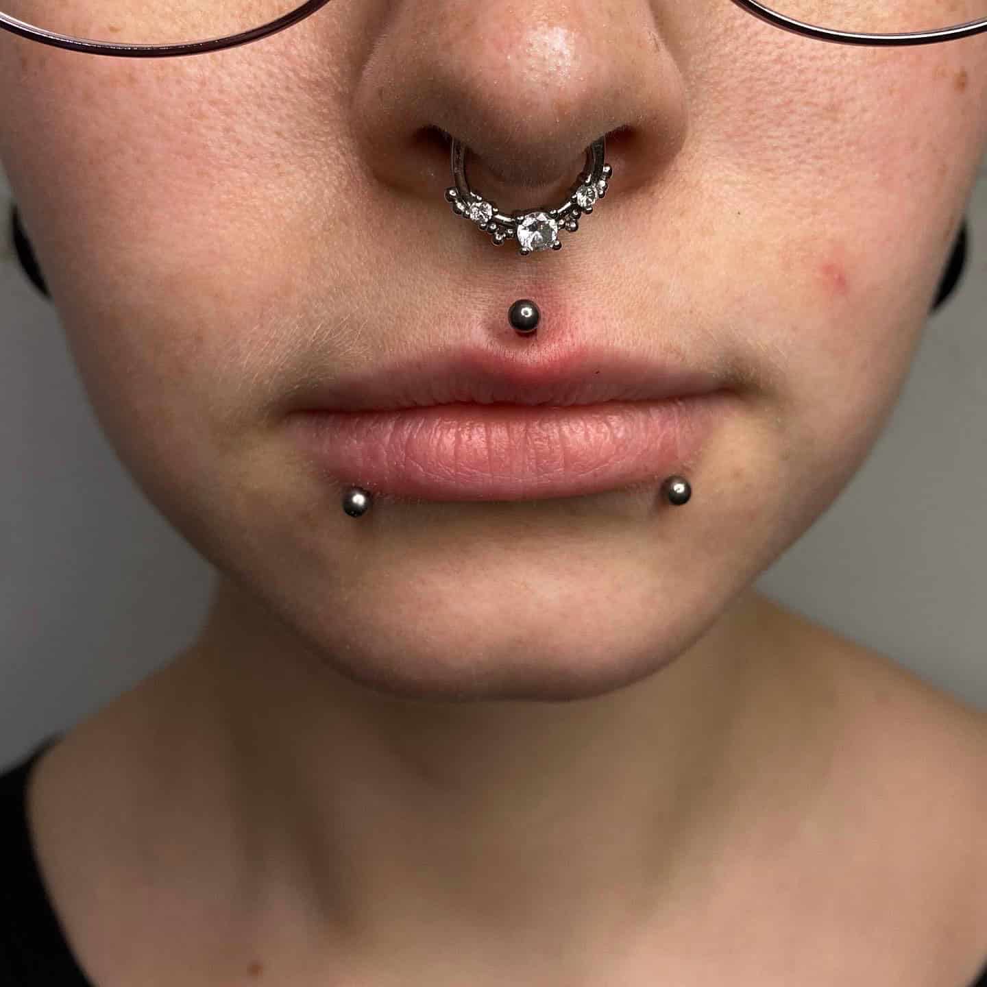 7sinstattoo.live Zbliżenie twarzy osoby z wieloma piercingami w studio tatuażu Poznań. Są to piercing przegrody nosowej z biżuterią, pionowy piercing wargi z kulką na końcu oraz dwa piercingi po każdej stronie dolnej wargi. Osoba ma na sobie okulary, a część jej szyi i koszuli jest widoczna. Nazwa produktu: 6107C23D-E30B-4A0D-80A2-0091C4B26C7C. Najlepsze studio tatuażu Seven Sins Tattoo