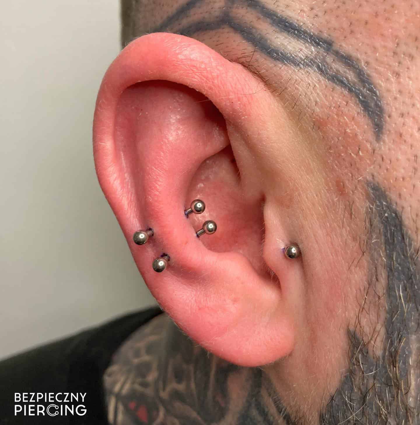 7sinstattoo.live Zbliżenie ucha osoby ozdobionego wieloma piercingami, z dwoma srebrnymi sztangami w wewnętrznej chrząstce i dwoma w zewnętrznej chrząstce. Tatuaże obejmujące ucho podkreślają kompetencje studia tatuażu Poznań. Tekst „BEZPIECZNY PIERCING” jest wyświetlany w lewym dolnym rogu. Nazwa produktu: 610D03E3-B222-4A7B-A344-19391237608F. Najlepsze studio tatuażu Seven Sins Tattoo