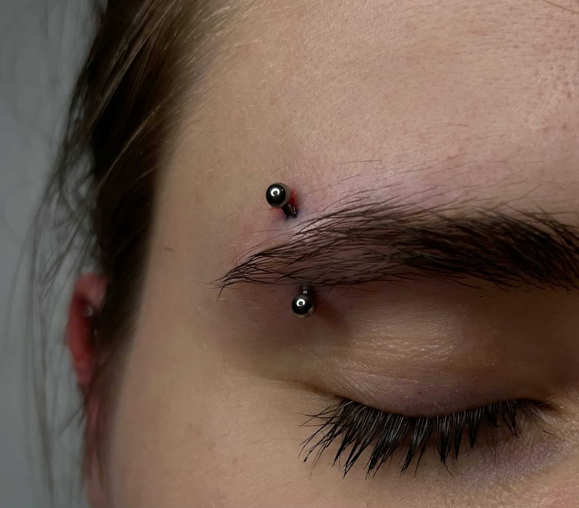 7sinstattoo.live Zbliżenie na zamknięte oko osoby pokazujące piercing brwi. Piercing składa się z pręta z czarnymi kulkami na każdym końcu, przewleczonego przez skórę nad brwią, z odrobiną zaczerwienienia wokół punktów wejścia i wyjścia. Uchwycone w *studio tatuażu Poznań*, oko i rzęsy są widoczne. Polecany produkt: 62A1954A-9358-4D49-AA56-91AC6E07C17E. Najlepsze studio tatuażu Seven Sins Tattoo