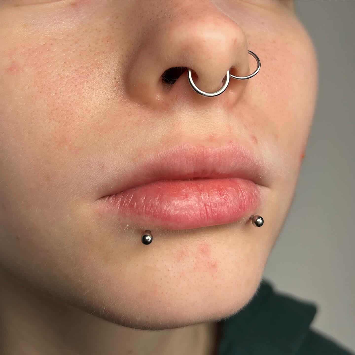 7sinstattoo.live Zbliżenie nosa i ust osoby, z dwoma srebrnymi pierścieniami przebijającymi przegrodę nosową i dwoma kolczykami z kulkami zdobiącymi dolną wargę. Skóra wydaje się lekko zarumieniona na neutralnym tle, prezentując misterne wykonanie studia tatuażu Poznań dla produktu 6AED672E-4427-4355-9726-21B278396364. Najlepsze studio tatuażu Seven Sins Tattoo