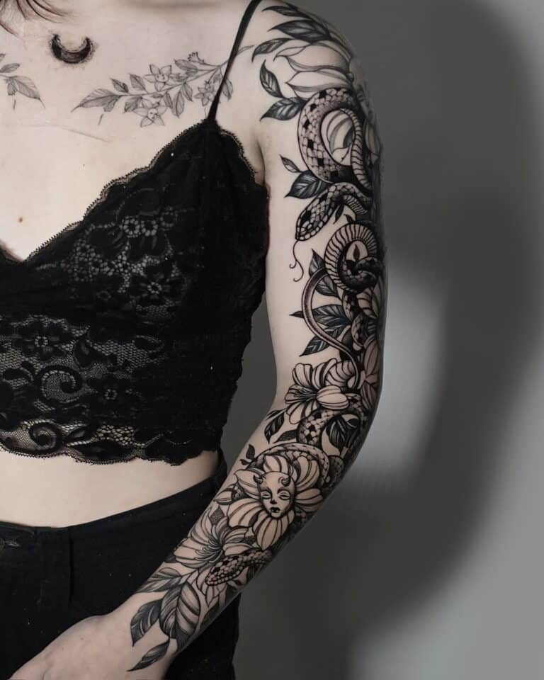 Osoba ubrana w misternie zaprojektowany czarny koronkowy top „7” i czarne spodnie z 7sinstattoo.live prezentuje szczegółowy tatuaż na lewym ramieniu. Tatuaż, wykonany w Seven Sins Tattoo Studio w Poznaniu, zawiera różne motywy kwiatowe, płynne wzory, półksiężyc na ramieniu i twarz kobiety płynnie zintegrowaną z dziełem sztuki. Dodatkowe tatuaże są również widoczne na klatce piersiowej.