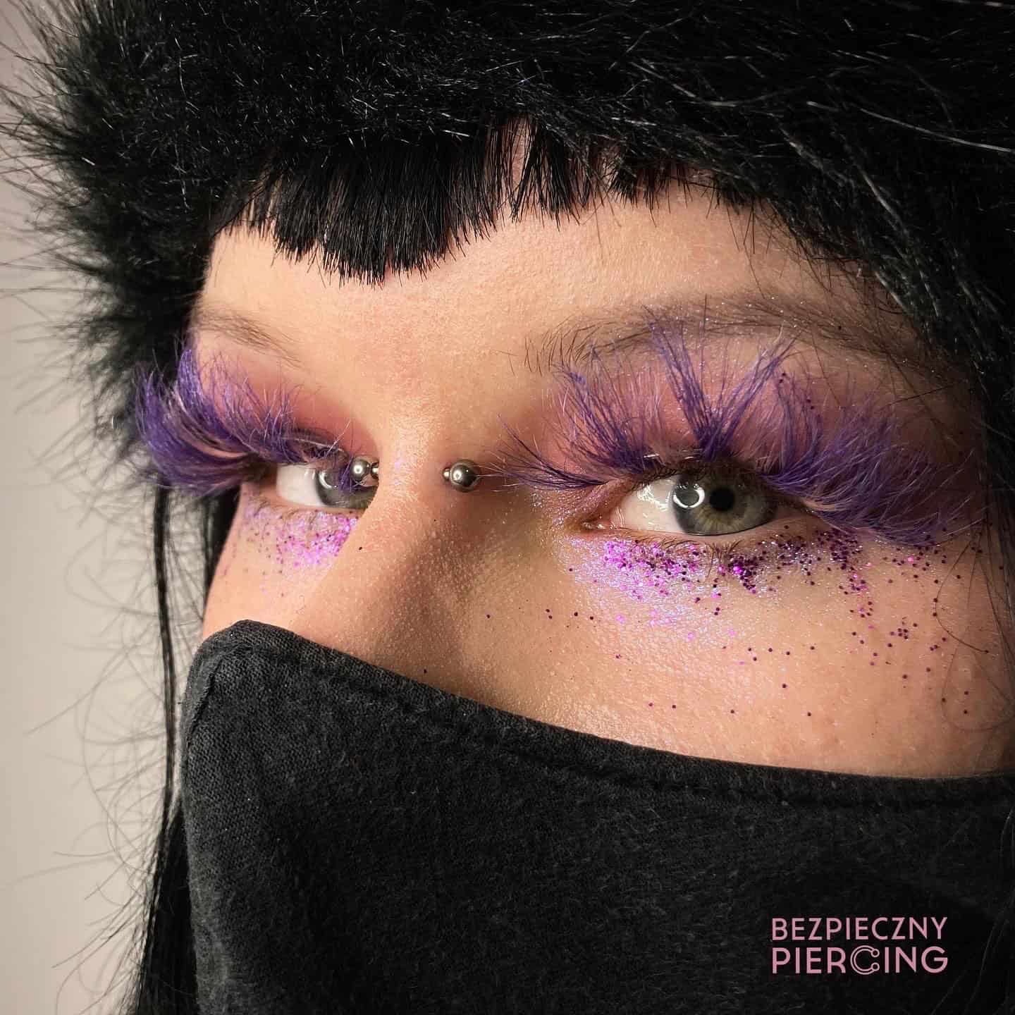 7sinstattoo.live Zbliżenie oczu osoby z misternymi fioletowymi, pierzastymi rzęsami i błyszczącym różowym cieniem do powiek, prezentujące mały metalowy piercing skóry nad grzbietem nosa. Nosi czarną maskę na twarzy i ma czarne włosy z grzywką. Tekst w prawym dolnym rogu mówi „BEZPIECZNY PIERCING - studio tatuaży Poznań”. Nazwa produktu: 79E5932F-E40F-48CF-A590-764DA0B0FDF1. Najlepsze studio tatuażu Seven Sins Tattoo