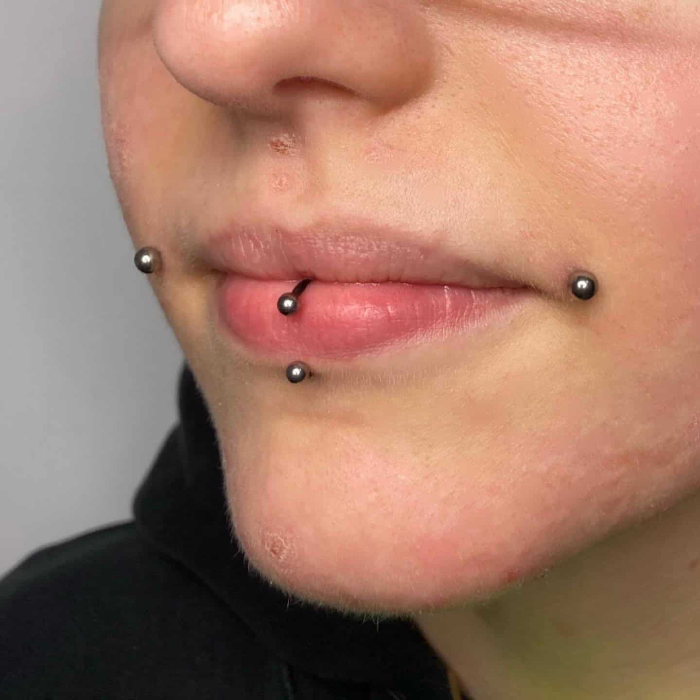 7sinstattoo.live Zbliżenie dolnej części twarzy osoby z trzema piercingami w pobliżu ust: dwa po każdej stronie dolnej wargi (piercingi typu snake bite) i jeden piercing w środku. Metalowe ćwieki łapią światło, gdy noszą czarny top, prawdopodobnie marki 8A2DC004-DD18-4575-B6CF-AB860A969D5C. Najlepsze studio tatuażu Seven Sins Tattoo