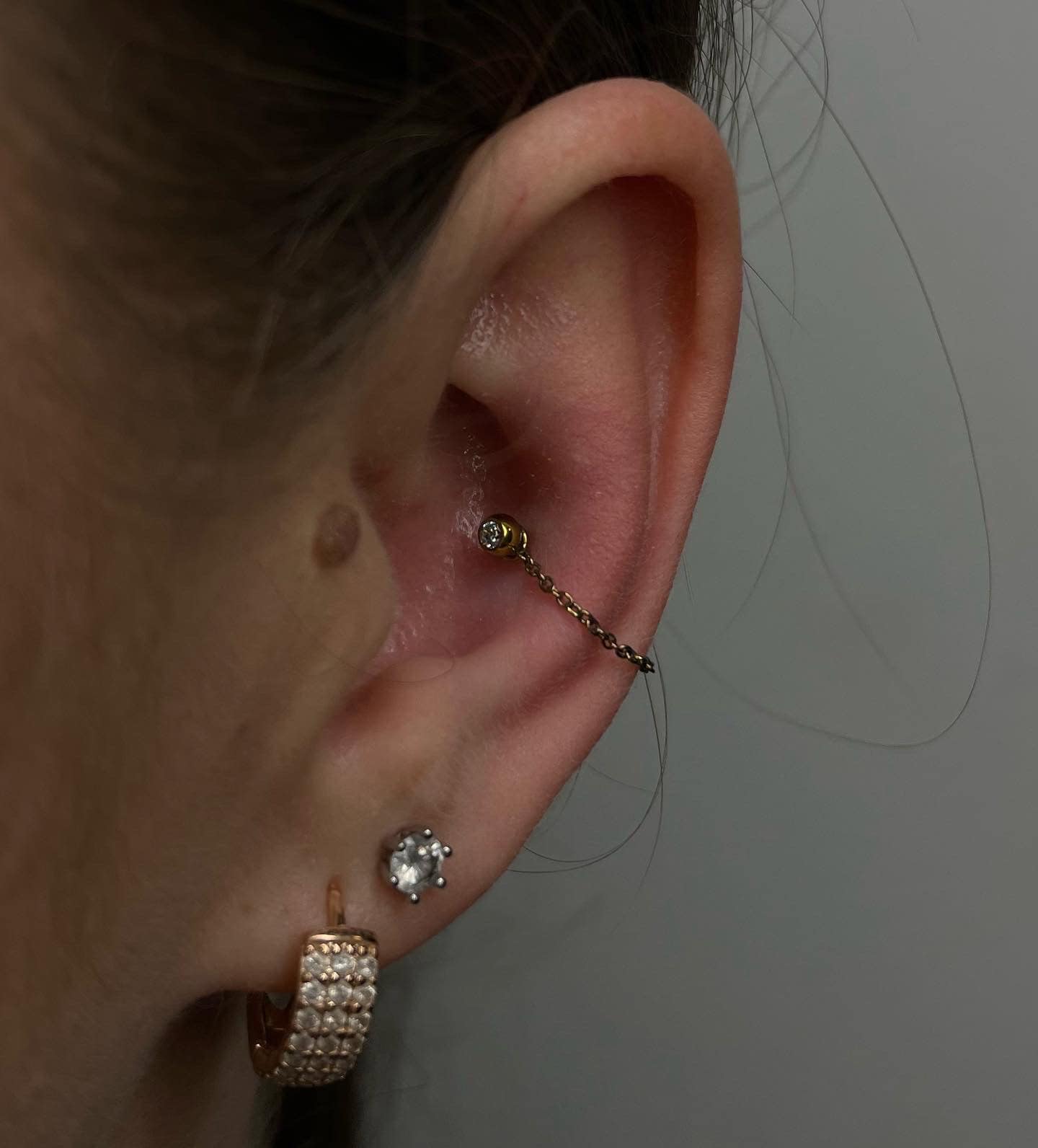 7sinstattoo.live Zbliżenie na ucho z kilkoma piercingami wykonanymi w najlepszym studiu tatuażu w Poznaniu. W uchu znajduje się piercing daith ozdobiony produktem 923914D9-67C9-4504-BC07-900D423CC528, złotym kolczykiem w kształcie kwiatu połączonym cienkim łańcuszkiem z piercingiem helix. Dodatkowo są dwa piercingi płatka ucha: jeden z błyszczącym diamentowym kolczykiem, a drugi z kolczykiem w kształcie kółka. Najlepsze studio tatuażu Seven Sins Tattoo