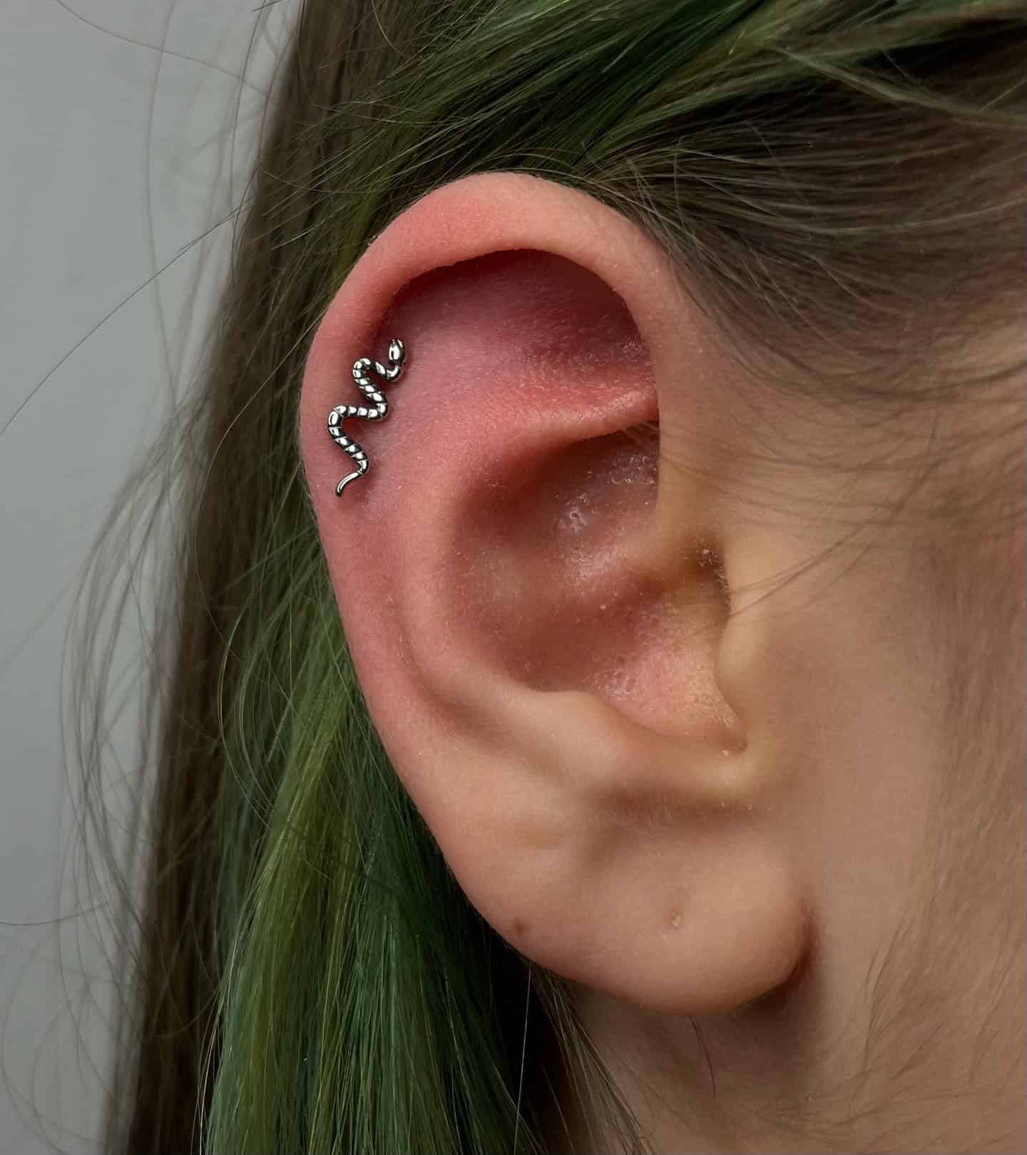 7sinstattoo.live Zbliżenie ucha z włosami w odcieniu zieleni, z kolczykiem w kształcie węża w górnej chrząstce. Srebrny kolczyk o nazwie 9556375F-F7B9-4AF0-B339-33D35431B21B jest ozdobiony małymi kamieniami szlachetnymi, co nadaje mu lśniący wygląd. Miłośnicy sztuki na ciele powinni sprawdzić najlepsze studio tatuażu Poznań, aby znaleźć najwyższej klasy wzory. Najlepsze studio tatuażu Seven Sins Tattoo