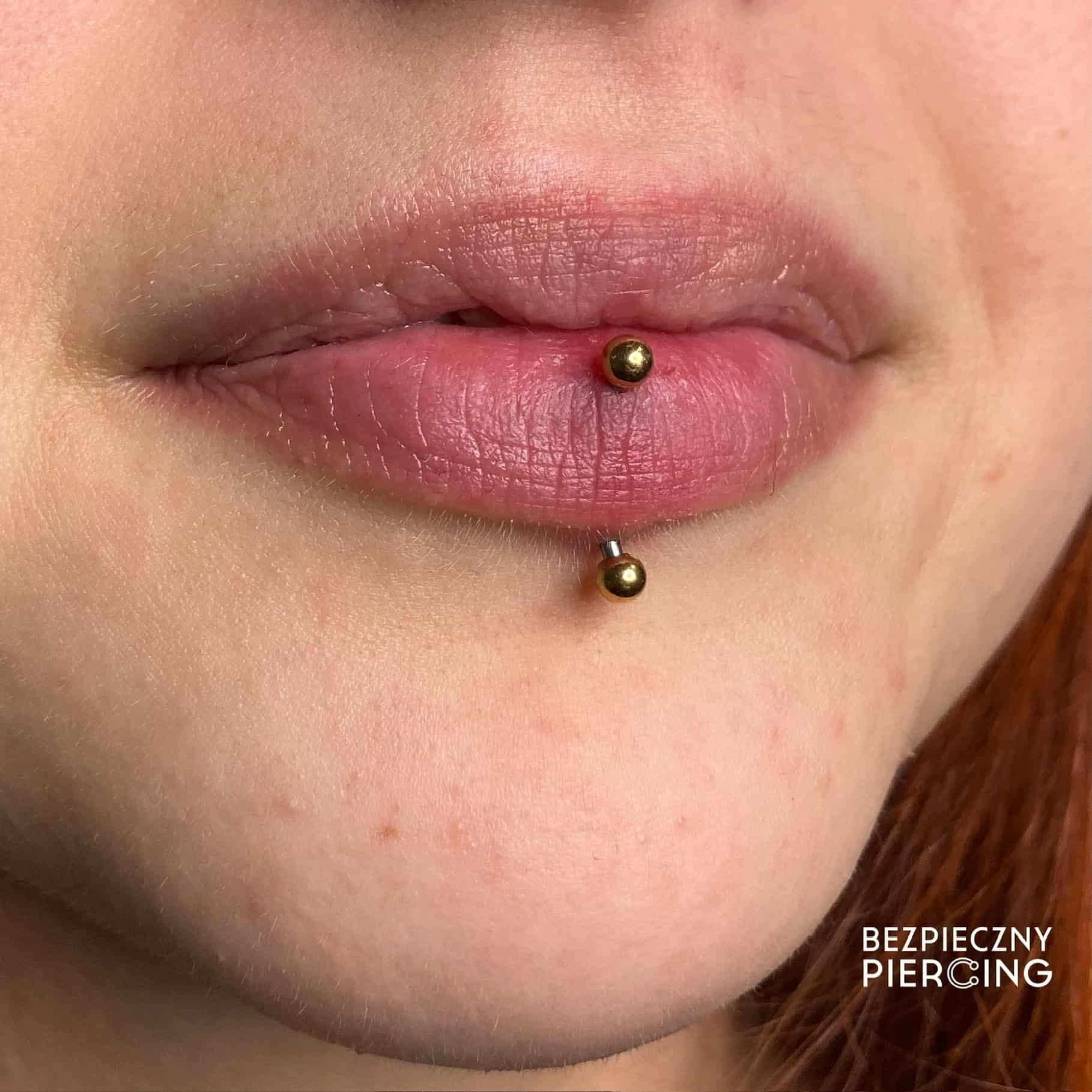 7sinstattoo.live Zbliżenie ust osoby z pionowym piercingiem labret z tatuaż Poznań. Piercing przedstawia "A78660F6-9590-489D-8431-C9EE2E017FA6" z dwoma złotymi kulkami na końcach, jedną nad i jedną pod dolną wargą. W prawym dolnym rogu pojawia się tekst "BEZPIECZNY PIERCING". Najlepsze studio tatuażu Seven Sins Tattoo