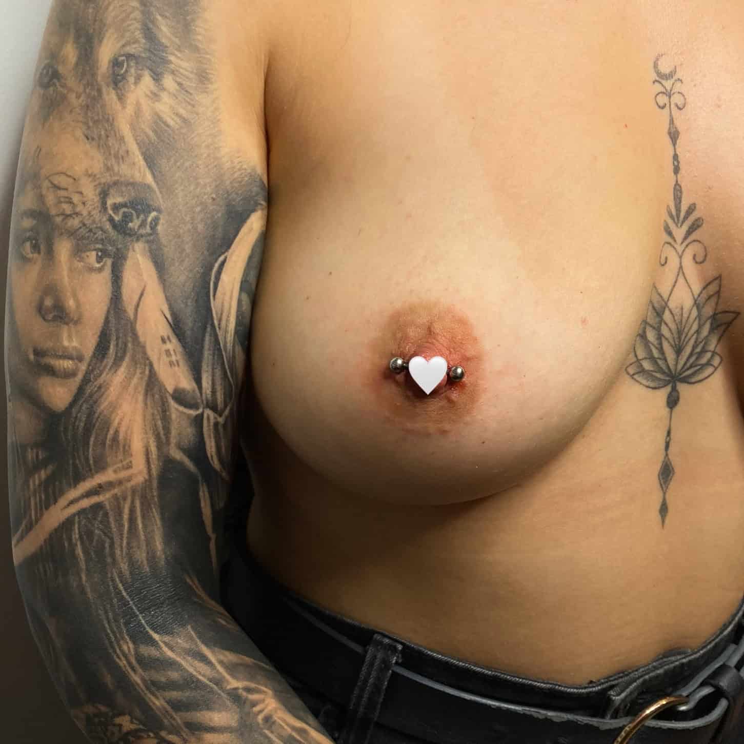 7sinstattoo.live Osoba z wytatuowanymi rękawami na ramionach, prawdopodobnie ze studia tatuaży Poznań, stoi topless w czarnych spodniach. Ma kolczyk w nosie i piercing w lewym sutku, który jest ozdobiony nakryciem w kształcie serca. Tatuaże mają skomplikowane wzory, w tym portret i obrazy zwierząt. Ten wizerunek jest zgodny z estetyką produktu AF6F23CD-AAA9-42A2-939C-58B01E200F34. Najlepsze studio tatuażu Seven Sins Tattoo