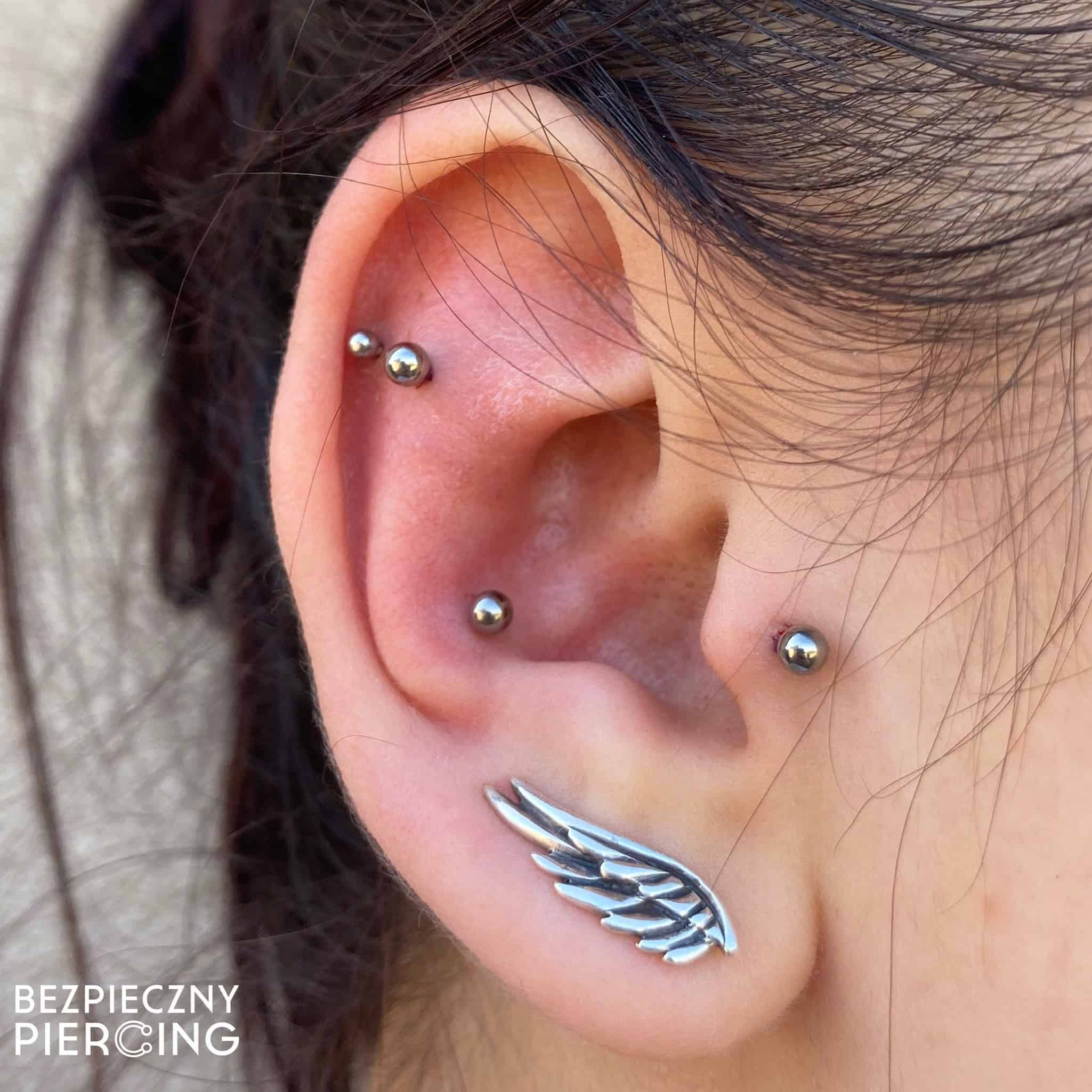 7sinstattoo.live Zbliżenie ucha zdobionego wieloma piercingami; płatek ucha szczyci się dużym kolczykiem w kształcie skrzydła anioła, a helisa, rook i tragus zdobią małe srebrne kulki. Na zdjęciu znajduje się znak wodny z napisem „Bezpieczny Piercing” pochodzący z renomowanego studia tatuażu Poznań. Najlepsze studio tatuażu Seven Sins Tattoo