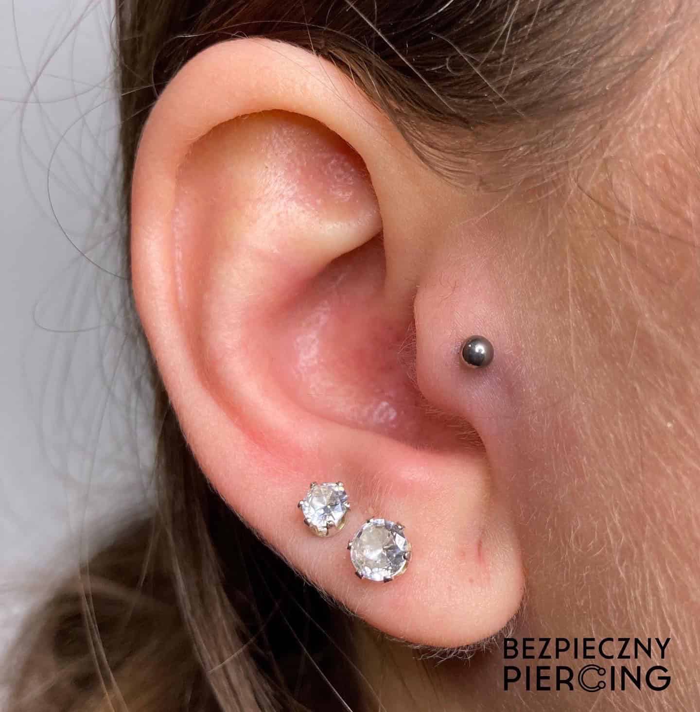 7sinstattoo.live Zbliżenie ucha osoby ozdobionego dwoma kolczykami z przezroczystymi kamieniami szlachetnymi w płatku ucha i małym srebrnym kolczykiem w kształcie kulki w chrząstce, prezentującym „Product BCBA5123-859E-4F98-BDB8-F9B35C5DDBF6”. Tło jest gładkie z napisem „BEZPIECZNY PIERCING” w prawym dolnym rogu, podkreślającym estetyczne doświadczenie Studia Tatuaże Poznań. Najlepsze studio tatuażu Seven Sins Tattoo
