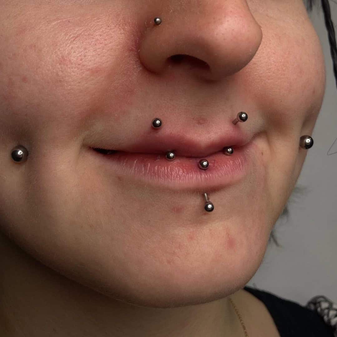 7sinstattoo.live Zbliżenie dolnej części twarzy osoby pokazujące wiele piercingów twarzy ozdobionych małą metalową biżuterią w kształcie kulek, w tym sześć piercingów na ustach i okolicach, takich jak nos i dołeczki. Osoba ma delikatny uśmiech, pokazując swój unikalny styl z CFEA723B-7BB9-43E0-B620-F338BF248910 z tatuaż Poznań. Najlepsze studio tatuażu Seven Sins Tattoo