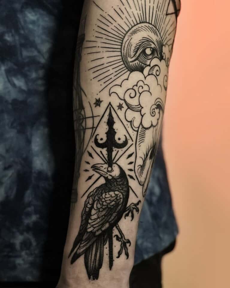 Zbliżenie tatuażu na przedramieniu z kolekcji DSC00032 7sinstattoo.live, przedstawiającego czarnego kruka siedzącego na gałęzi, z geometrycznym wzorem przypominającym włócznię nad głową. Wokół kruka znajdują się chmury, półksiężyc i skomplikowany wzór linii tworzący wzór oka i rozbłysku słońca. Osoba ma na sobie ciemnoniebieską koszulę.