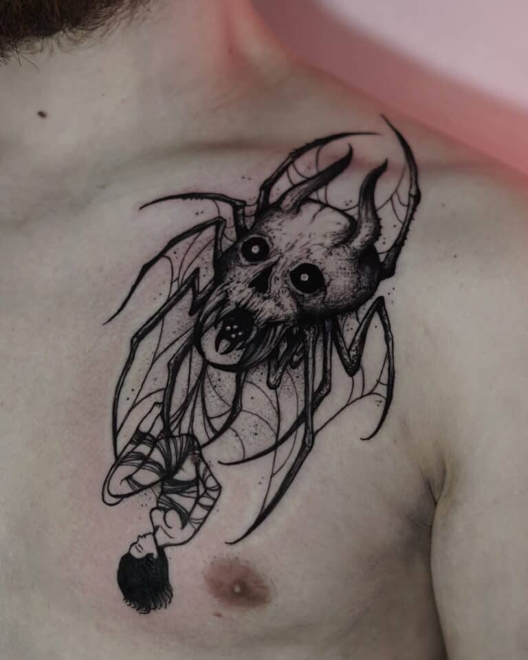Czarno-biały tatuaż na klatce piersiowej człowieka, misternie wykonany przez renomowane studio 7sinstattoo.live w Seven Sins Tattoo w Poznaniu, przedstawia groźną czaszkę z nietoperzowymi skrzydłami, kłami i ciemnymi oczami. Wzór obejmuje również wiszącą do góry nogami postać uosabiającą mroczną i gotycką esencję. To arcydzieło uchwycono na zdjęciu DSC00325.