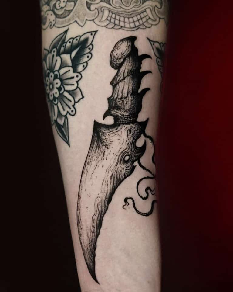 Czarno-biały tatuaż na przedramieniu przedstawia misternie zaprojektowany „7sinstattoo.live DSC00622 (1)” z Seven Sins Tattoo Studio w Poznaniu. Przedstawia zakrzywiony, ozdobny sztylet z rękojeścią ozdobioną szczegółowymi wzorami przypominającymi wielookie stworzenie z małymi mackami. Otaczające motywy kwiatowe wzmacniają ogólną kompozycję artystyczną stworzoną przez utalentowanego tatuażystę z Poznania.