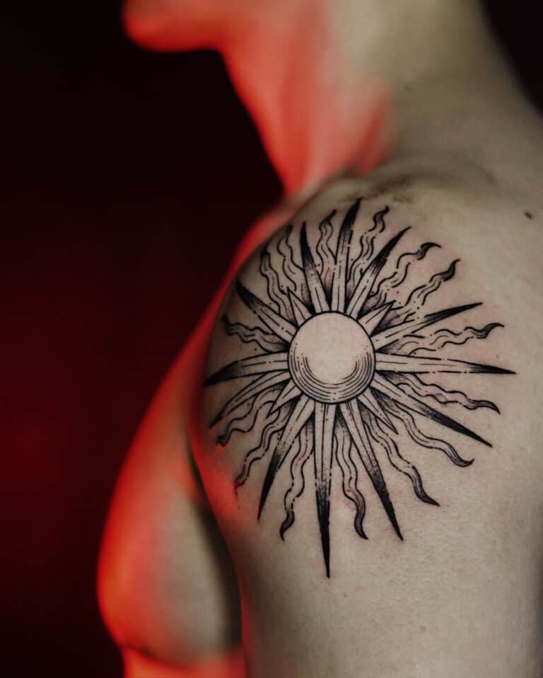 Czarno-biały tatuaż przedstawiający słońce ze stylizowanymi promieniami na górnej części ramienia, wykonany przez utalentowanych artystów z Seven Sins Tattoo w Poznaniu. Zdjęcie zatytułowane DSC00622 z 7sinstattoo.live zostało zrobione z boku i prezentuje misterny wzór na słabo oświetlonym tle z czerwonymi odcieniami, które podkreślają kunszt jednego z najlepszych poznańskich studiów.