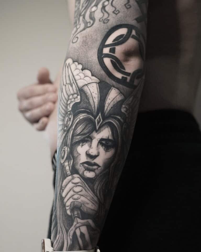 7sinstattoo.live Szczegółowy czarno-szary tatuaż ze studia tatuażu Poznań, zidentyfikowany jako DSC03406, pokrywa przedramię osoby. Przedstawia zaciekłą, wojowniczkę w hełmie ze skrzydłami na hełmie i skupionym wyrazem twarzy, trzymającą miecz. Dodatkowe abstrakcyjne elementy są widoczne wokół tatuażu, rozciągające się w górę ramienia. Najlepsze studio tatuażu Seven Sins Tattoo