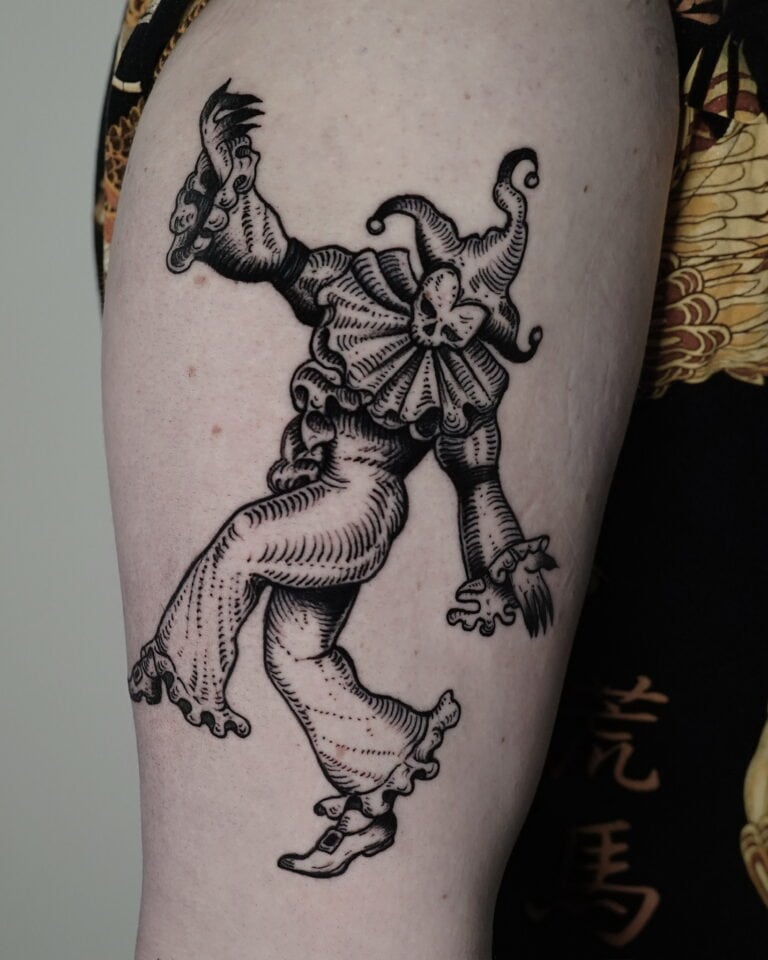7sinstattoo.live Zbliżenie na ramię osoby pokazuje szczegółowy czarno-szary tatuaż błazna w klasycznym stroju z falbaniastym kołnierzem, kapeluszem i spiczastymi butami. Błazen jest przedstawiony w animowanej, żywej pozie, emanującej kaprysem. Uchwycony jako DSC04234 w tatuaż Poznań, osoba ma na sobie ciemne, wzorzyste ubranie. Najlepsze studio tatuażu Seven Sins Tattoo