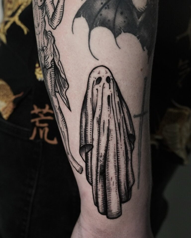 7sinstattoo.live Zbliżenie tatuażu z DSC04248, wykonanego w Najlepszym Studio Tatuażu Poznań, przedstawia czarno-białą, upiorną postać udrapowaną w zwiewnym prześcieradle z otworami na oczy. Wokół centralnego fragmentu znajdują się inne tatuaże o ciemnej tematyce, w tym szkieletowa postać i skrzydła nietoperza. Strój noszącego jest ozdobiony azjatyckimi wzorami postaci. Najlepsze studio tatuażu Seven Sins Tattoo