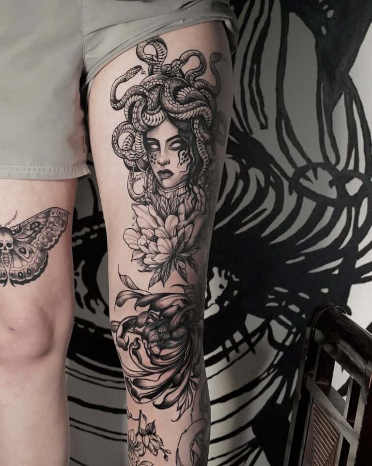 7sinstattoo.live Wytatuowana noga z dużą głową Meduzy z włosem węża, różą i innymi skomplikowanymi wzorami, takimi jak ćma i krab, rozciąga się od połowy uda do kolana. Szczegółowe monochromatyczne dzieło sztuki wyróżnia się na tle czarnego muralu, wszystko fachowo wykonane w studiu DSC04304-восстановлено w Poznaniu. Najlepsze studio tatuażu Seven Sins Tattoo