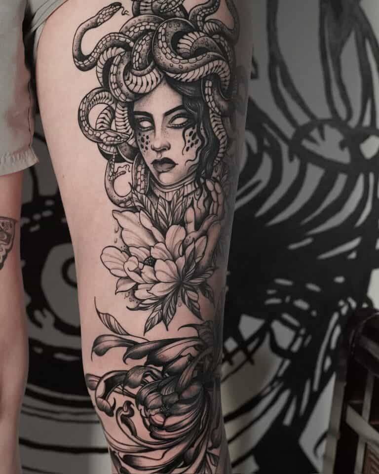 7sinstattoo.live Czarno-biały tatuaż na górnej części uda osoby przedstawiający głowę Meduzy z włosami węża, otoczony kwiatowymi wzorami i skomplikowanymi wzorami. Szczegółowe dzieło sztuki, wykonane w najlepszym studio tatuażu w Poznaniu, obejmuje znaczną część nogi. | Nazwa produktu: DSC04312 Najlepsze studio tatuażu Seven Sins Tattoo