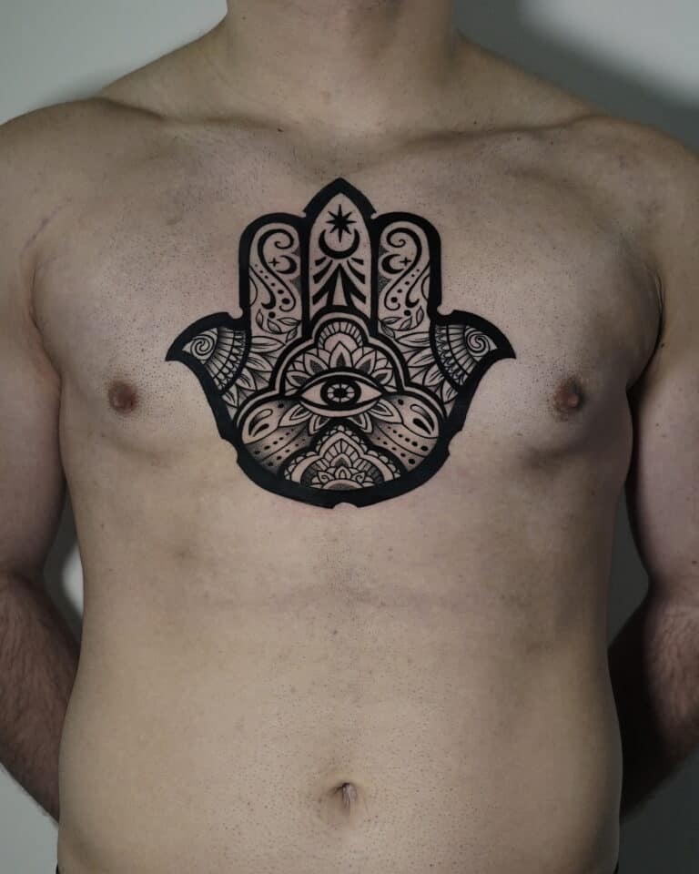 7sinstattoo.live Osoba z dużym, misternym czarnym tatuażem symbolu Hamsa na piersi stoi na prostym tle. Wzór charakteryzuje się ozdobnymi wzorami i obejmuje oko w środku dłoni, rozciągające się na górną część klatki piersiowej. Ta oszałamiająca praca została uchwycona w produkcie DSC04471 w studio tatuażu Poznań. Najlepsze studio tatuażu Seven Sins Tattoo