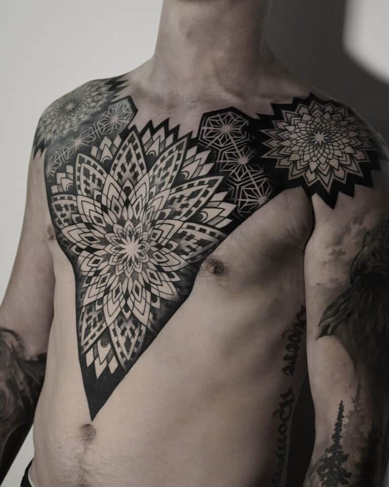 7sinstattoo.live Osoba prezentuje misterny i rozległy czarno-szary tatuaż mandali, który rozciąga się na klatce piersiowej i ramionach, z dodatkowymi mniejszymi tatuażami zdobiącymi jej ramię. Oświetlenie podkreśla szczegółowy projekt i cieniowanie widocznego tatuażu na klatce piersiowej, umiejętnie wykonanego w tatuaż Poznań, jak uchwycono w produkcie DSC04850. Najlepsze studio tatuażu Seven Sins Tattoo