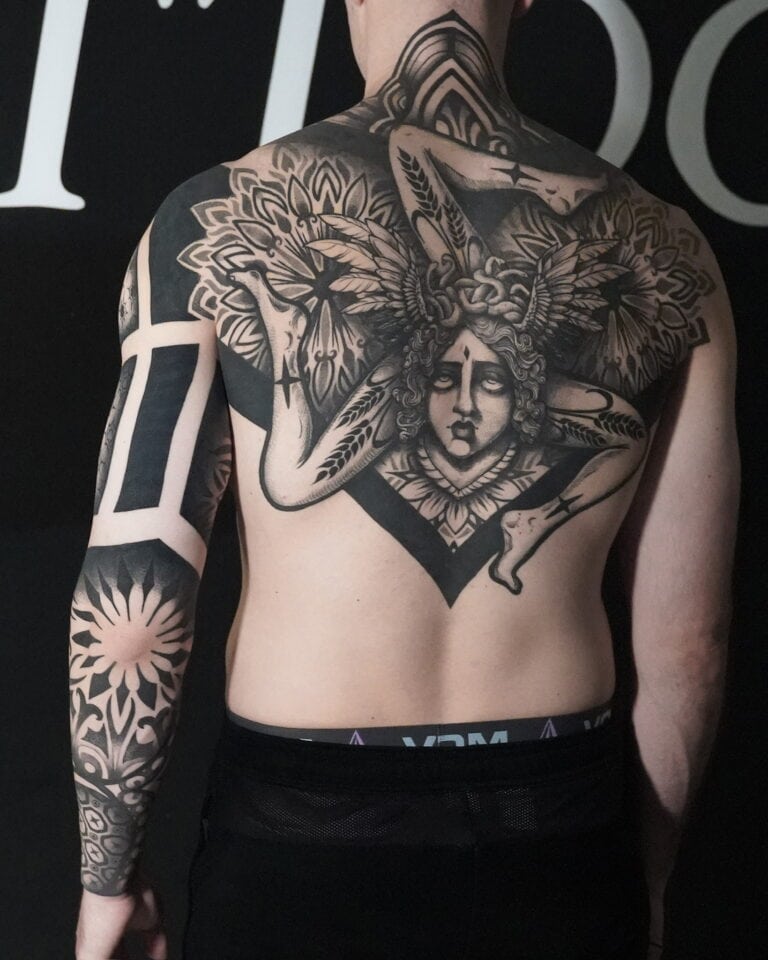 7sinstattoo.live Na plecach osoby znajduje się szczegółowy czarno-szary tatuaż z geometrycznym wzorem i postacią z misternymi skrzydłami oraz ozdobnym nakryciem głowy, stworzony w najlepszym studio tatuażu w Poznaniu. Osoba ta ma również abstrakcyjne i kwiatowe tatuaże na prawym ramieniu. Więcej szczegółów można znaleźć w nazwie produktu: DSC05897. Najlepsze studio tatuażu Seven Sins Tattoo