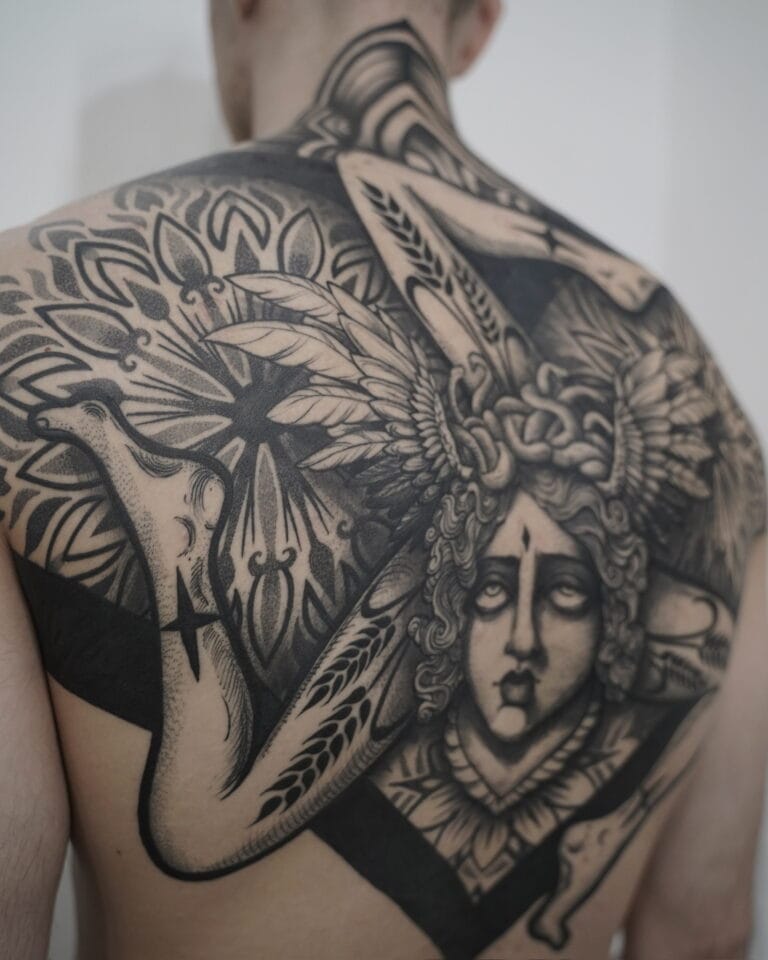 7sinstattoo.live Na plecach osoby znajduje się DSC07467, misterny czarno-szary tatuaż. Skomplikowany projekt przedstawia centralną postać z szczegółową twarzą i włosami, otoczoną symetrycznymi, abstrakcyjnymi i geometrycznymi wzorami, które rozciągają się na górnej części pleców i ramionach. To arcydzieło zostało wykonane w jednym z najlepszych studiów tatuażu w Poznaniu. Najlepsze studio tatuażu Seven Sins Tattoo