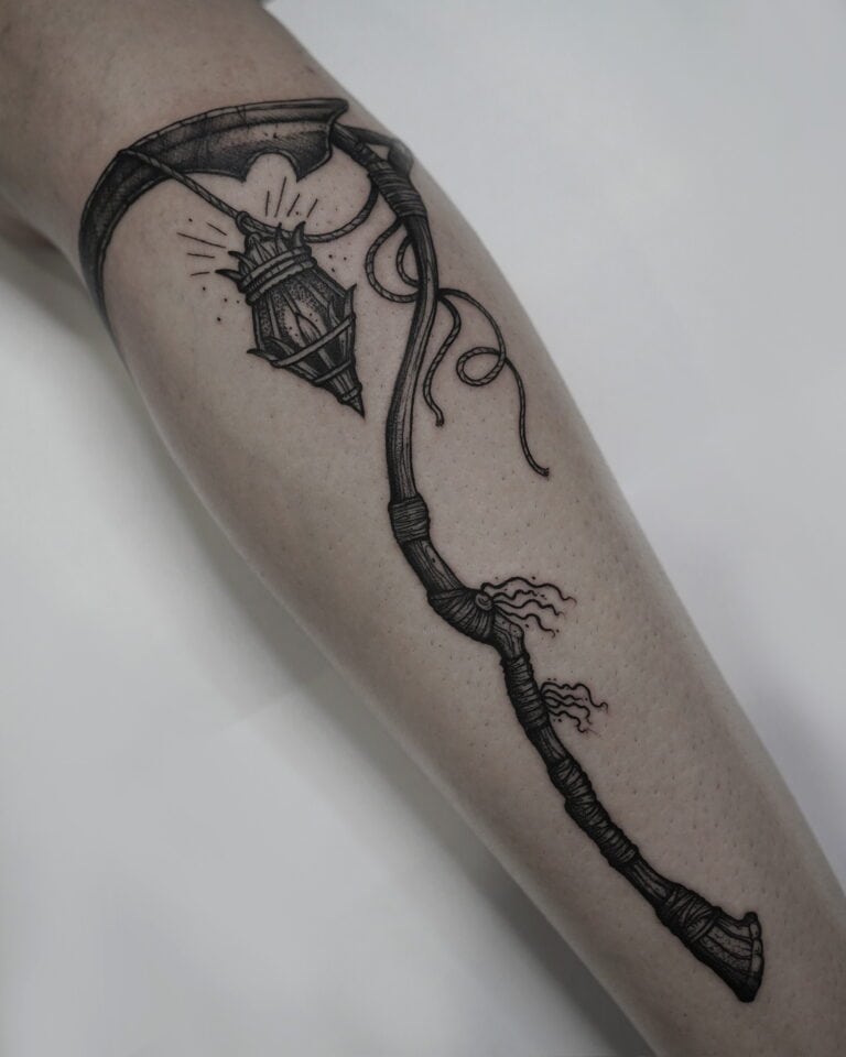 7sinstattoo.live Tatuaż DSC07641 na ramieniu przedstawia szczegółowo odwzorowaną czarno-szarą latarnię uliczną zawieszoną na zakrzywionym, zwiniętym słupie z małym toporem na końcu. Lampa emituje świecące promienie światła. Słup zdobią misterne wiązania i wstążki, mistrzowsko wykonane przez najlepsze studio tatuażu w Poznaniu. Najlepsze studio tatuażu Seven Sins Tattoo
