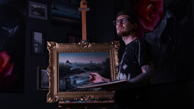 7sinstattoo.live Osoba w okularach i z tatuażami maluje na płótnie umieszczonym na sztaludze. Pomieszczenie przypominające artystyczne studio tatuażu Poznań jest słabo oświetlone i ozdobione oprawionymi dziełami sztuki. Pędzel artysty dotyka obrazu krajobrazowego przedstawiającego scenę rzeki uchwyconą na oszałamiającym zdjęciu DSC09499. Najlepsze studio tatuażu Seven Sins Tattoo