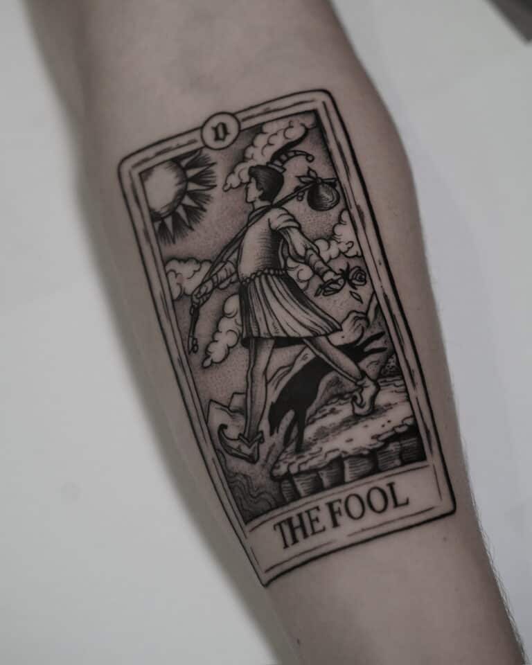 7sinstattoo.live Szczegółowy czarno-biały tatuaż karty Tarota „Głupiec” na przedramieniu, uchwycony na zdjęciu produktu DSC09590 z najlepszego studia tatuażu Poznań. Wzór przedstawia osobę w stroju błazna schodzącą z klifu, trzymającą mały woreczek na kiju na ramieniu, ze słońcem i chmurami w tle. Najlepsze studio tatuażu Seven Sins Tattoo