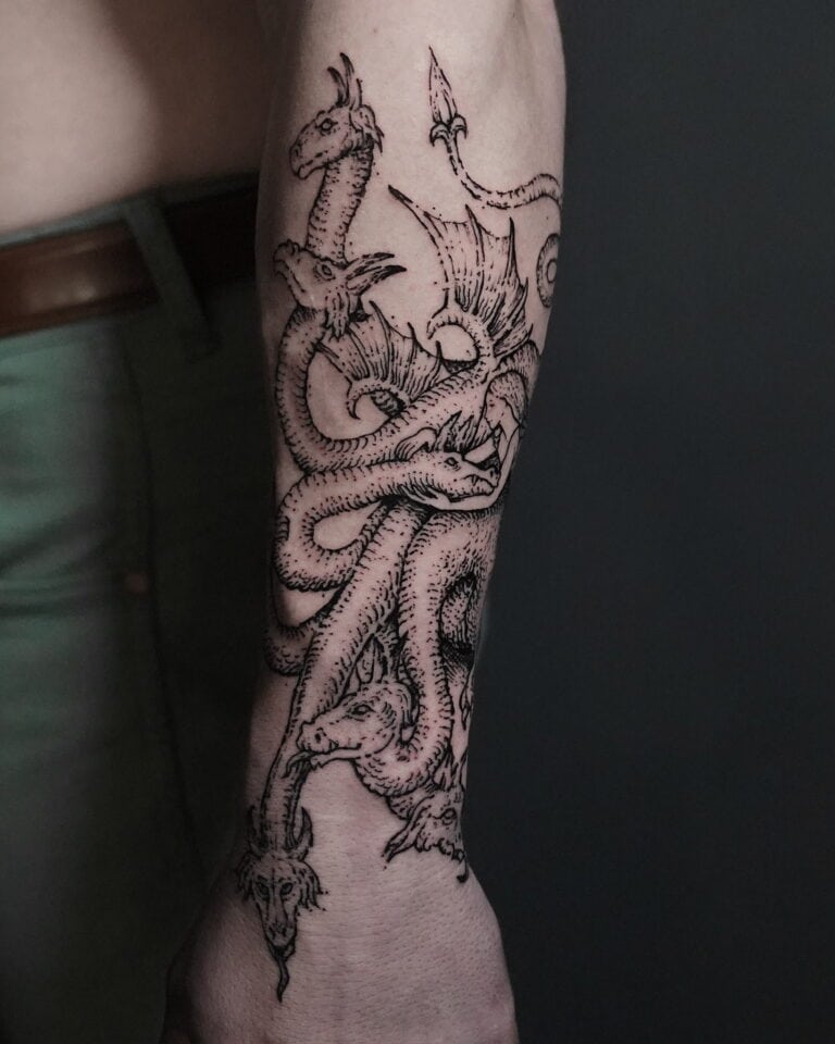 7sinstattoo.live Zbliżenie na przedramię osoby, na którym widoczny jest skomplikowany tatuaż wielogłowego smoka, wykonany przez utalentowanego artystę z najlepszego studia tatuażu w Poznaniu. Szczegółowa praca liniowa ukazuje głowy smoków warczące i splatające się wzdłuż przedramienia. Osoba, zidentyfikowana jako DSC09812, ma na sobie zielone spodnie. Najlepsze studio tatuażu Seven Sins Tattoo