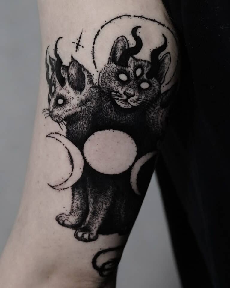 7sinstattoo.live Oszałamiający tatuaż zatytułowany „DSC09934”, stworzony w najlepszym studio tatuażu Poznań, przedstawia dwugłowego kota z trzema oczami i rogami. Obie głowy mistycznego stworzenia mają świecące oczy i są otoczone elementami niebieskimi, w tym fazami księżyca. Ta czarno-biała grafika zdobi ramię osoby na ciemnym tle. Najlepsze studio tatuażu Seven Sins Tattoo