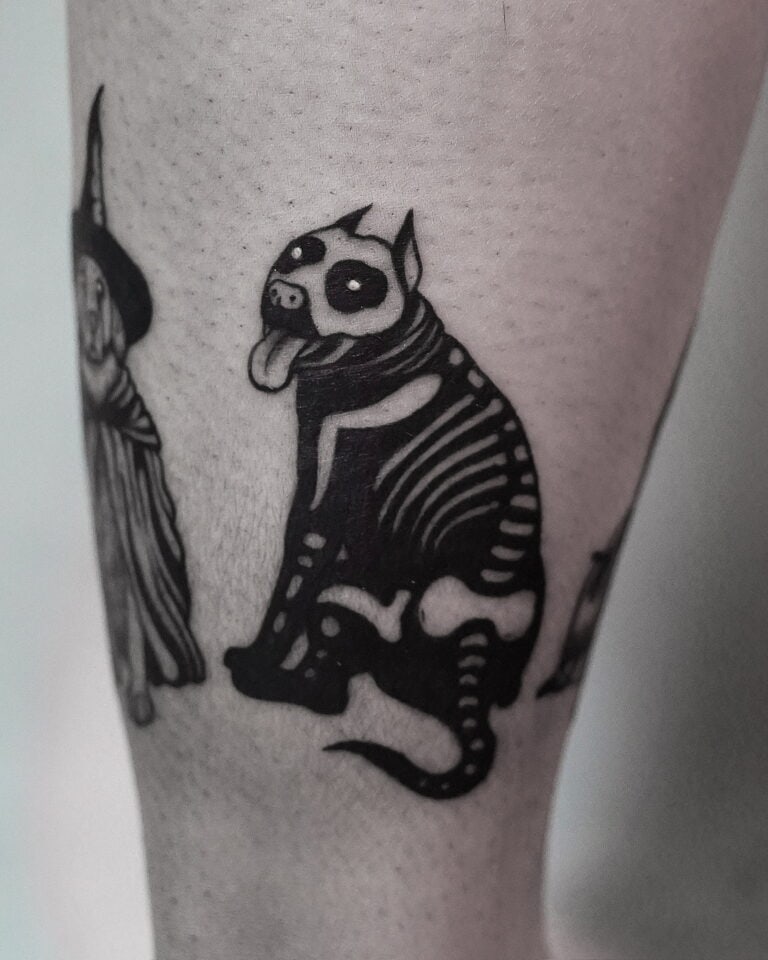 7sinstattoo.live Tatuaż na nodze osoby przedstawiający czarno-białego stylizowanego psa o szkieletowych rysach i wystającym języku, obok częściowego obrazu postaci w szacie i kapeluszu. Projekt utrzymuje mroczny, kapryśny motyw i został wykonany przez artystę z najlepszego studia tatuażu Poznań. Ta praca jest identyfikowana jako DSC09947. Najlepsze studio tatuażu Seven Sins Tattoo